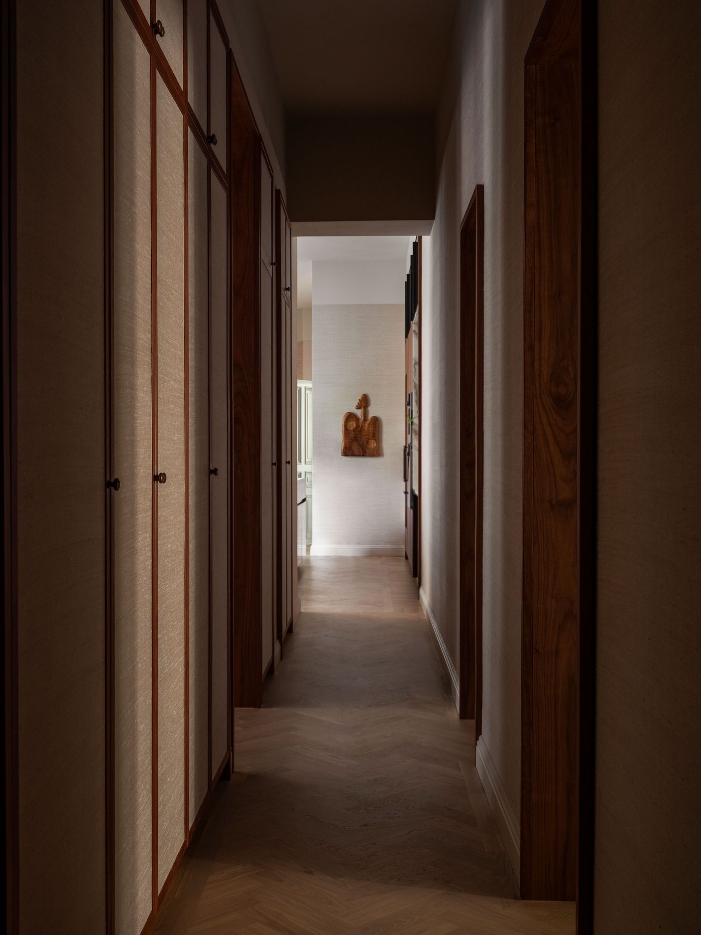 A warmly lit hallway featuring wooden cabinets, textured walls, and a decorative artwork, leading to an illuminated space. A warmly lit hallway featuring wooden cabinets, textured walls, and a decorative artwork, leading to an illuminated space.
