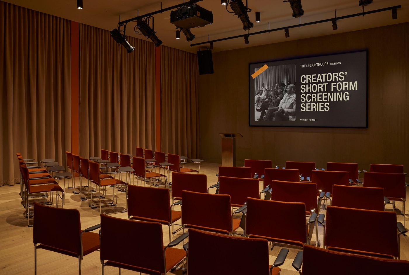 A modern screening room with orange chairs arranged in rows, featuring a large screen displaying "Creators' Short Form Screening Series." A modern screening room with orange chairs arranged in rows, featuring a large screen displaying "Creators' Short Form Screening Series."