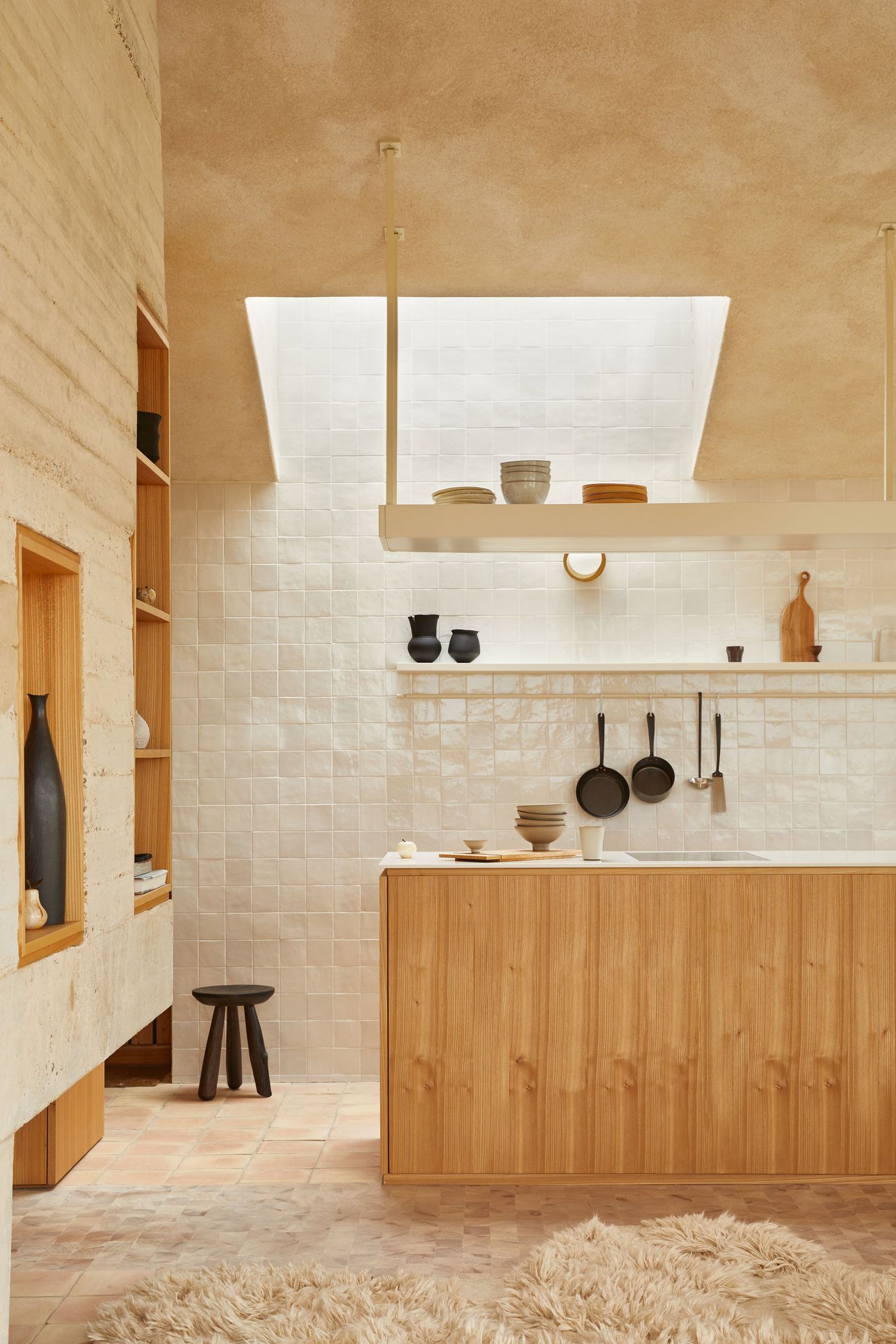 The kitchen of The Maker's Barn by Hutch Design exudes minimalist elegance with a focus on natural materials. Pale, textured square tiles create a subtle backdrop to light wooden cabinetry and open shelving, while black hanging pots add contrast. Natural light streams from a large skylight, enhancing the warm tones of the brick flooring and soft textures, reflecting a functional yet artful space. The kitchen of The Maker's Barn by Hutch Design exudes minimalist elegance with a focus on natural materials. Pale, textured square tiles create a subtle backdrop to light wooden cabinetry and open shelving, while black hanging pots add contrast. Natural light streams from a large skylight, enhancing the warm tones of the brick flooring and soft textures, reflecting a functional yet artful space.