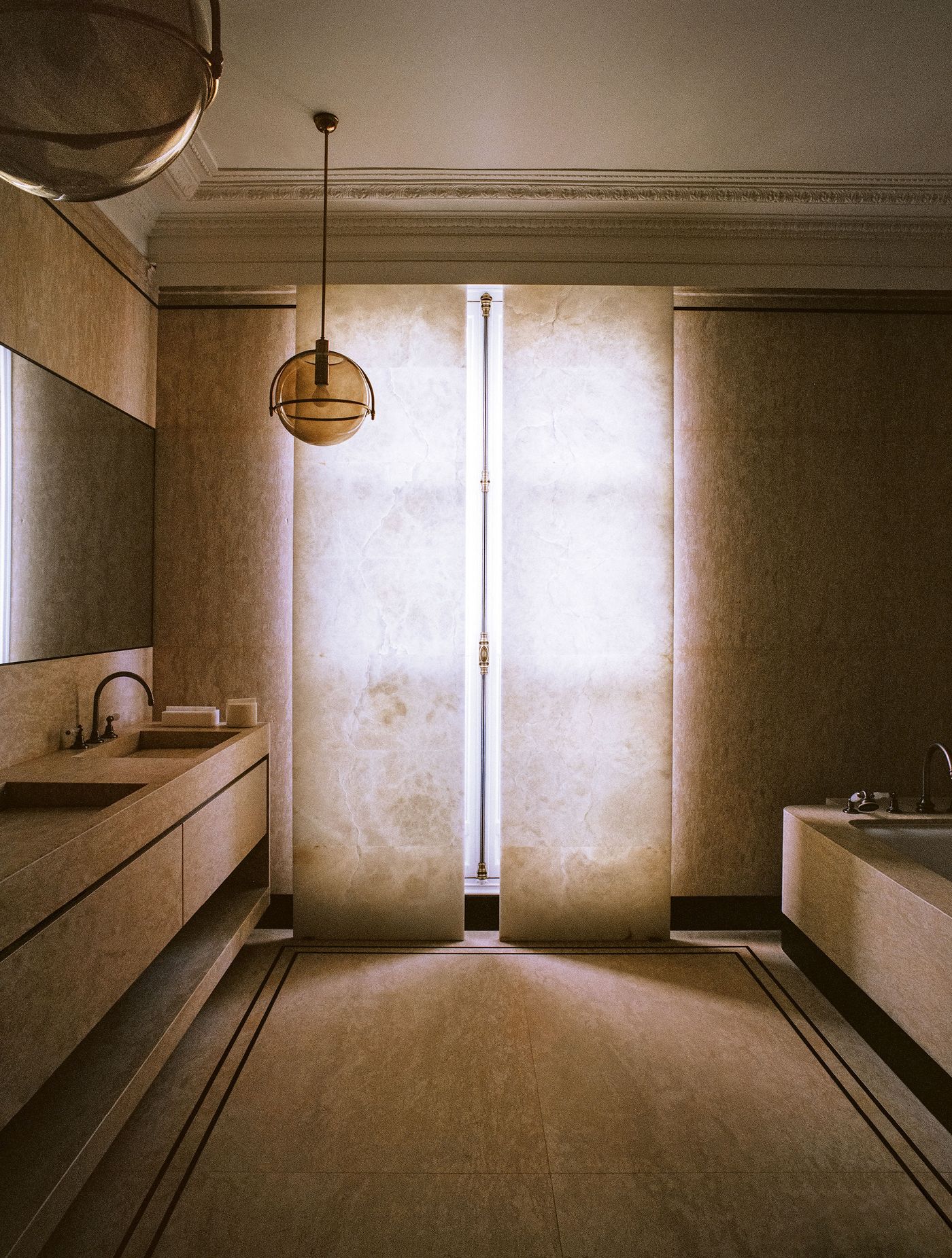 An opulent bathroom, entirely clad in light-toned natural stone, creating a monochromatic and serene atmosphere. Two large, back-lit translucent panels serve as a striking feature, complementing the minimalist double vanity and the freestanding bathtub, all illuminated by a delicate pendant light. An opulent bathroom, entirely clad in light-toned natural stone, creating a monochromatic and serene atmosphere. Two large, back-lit translucent panels serve as a striking feature, complementing the minimalist double vanity and the freestanding bathtub, all illuminated by a delicate pendant light.