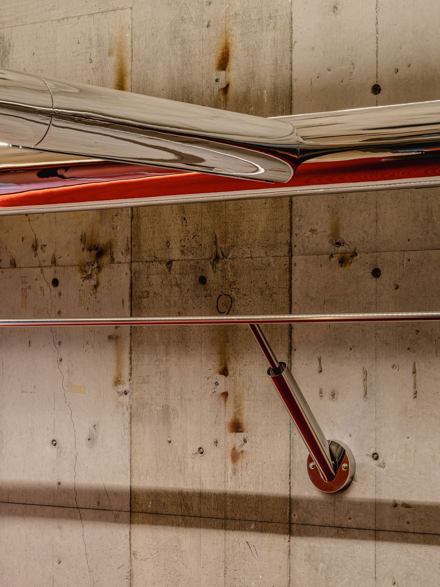 A sculptural junction in the chrome pipework folds and intersects against raw concrete, its highly reflective surface picking up flashes of red from the store. Below, a slender clothing rail is anchored by an angled polished support, turning functional retail infrastructure into a precise graphic composition.