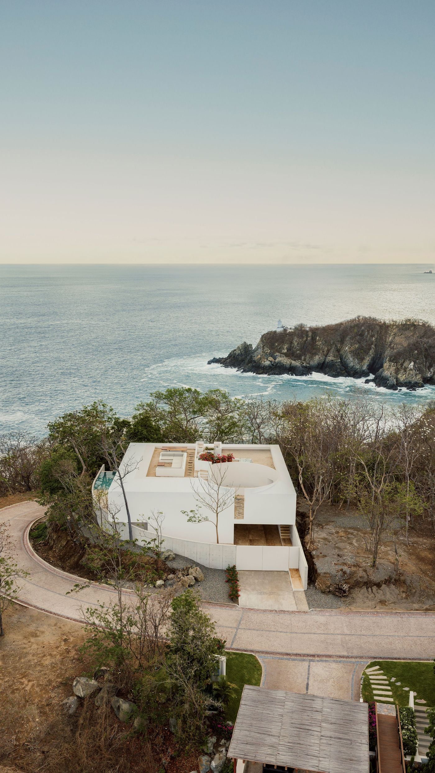 Seen from inland, Casa Malonic presents a closed white mass to the street, its exposed-concrete wall curving with the road towards a recessed entrance stair. Beyond the rooftop lounge and circular courtyard, a rocky outcrop crowned by a small lighthouse juts into a still, pastel-hued Pacific. Seen from inland, Casa Malonic presents a closed white mass to the street, its exposed-concrete wall curving with the road towards a recessed entrance stair. Beyond the rooftop lounge and circular courtyard, a rocky outcrop crowned by a small lighthouse juts into a still, pastel-hued Pacific.