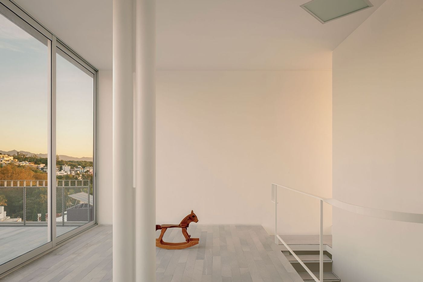 On Casa Tlaloc’s upper level, pale surfaces, slender white columns and expansive glazing create an airy, almost abstract interior. A small wooden rocking horse provides a warm, playful counterpoint, while the stair void and curved balustrade reveal the vertical connection between floors and sunset views extend across the landscape outside. On Casa Tlaloc’s upper level, pale surfaces, slender white columns and expansive glazing create an airy, almost abstract interior. A small wooden rocking horse provides a warm, playful counterpoint, while the stair void and curved balustrade reveal the vertical connection between floors and sunset views extend across the landscape outside.