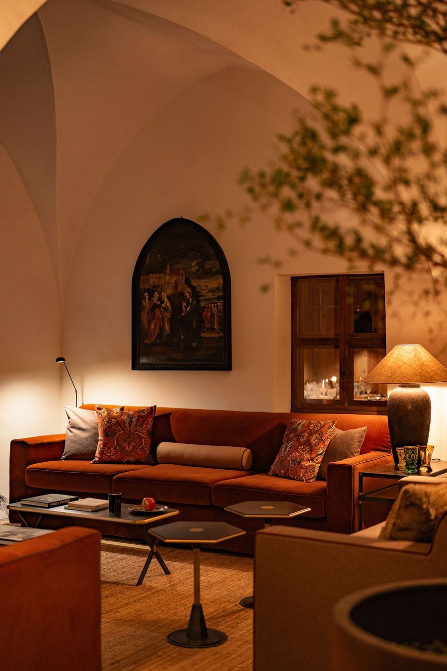 Pale groin vaults arch over a lounge at Castel Badia, where a rust velvet sofa with embroidered cushions sits beneath a pointed-arch painting of a biblical scene set before a hilltop castle. An interior window reveals a candlelit dining room, flanked by a stoneware lamp, coloured glass beakers and hexagonal side tables. Pale groin vaults arch over a lounge at Castel Badia, where a rust velvet sofa with embroidered cushions sits beneath a pointed-arch painting of a biblical scene set before a hilltop castle. An interior window reveals a candlelit dining room, flanked by a stoneware lamp, coloured glass beakers and hexagonal side tables.
