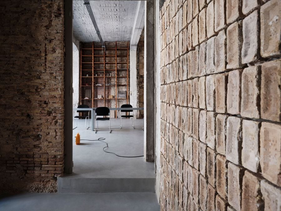 A narrow passage framed by exposed brick leads into Castor’s ground-floor workspace, where simple black chairs and metal tables sit against the continuous grey floor. In the background, a tall timber shelving unit introduces warmth amid the building’s raw brick, stone and concrete fabric.
