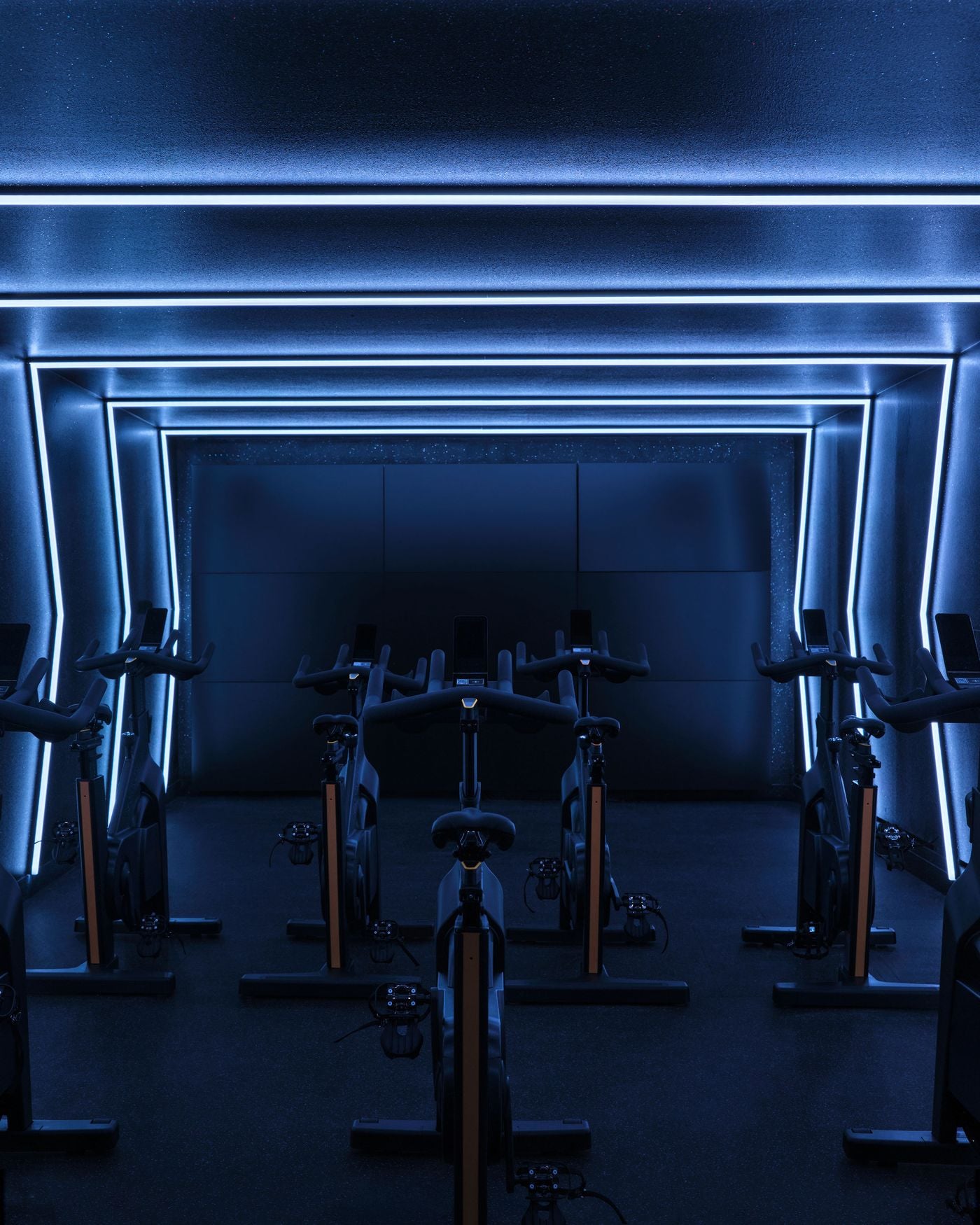 The cycling studio shifts into a darker, nightclub-like register, with rows of stationary bikes set against deep blue walls and ceiling. Continuous LED lines trace a series of nested rectangular frames around the room, amplifying depth and giving the space an immersive, high-energy atmosphere. The cycling studio shifts into a darker, nightclub-like register, with rows of stationary bikes set against deep blue walls and ceiling. Continuous LED lines trace a series of nested rectangular frames around the room, amplifying depth and giving the space an immersive, high-energy atmosphere.