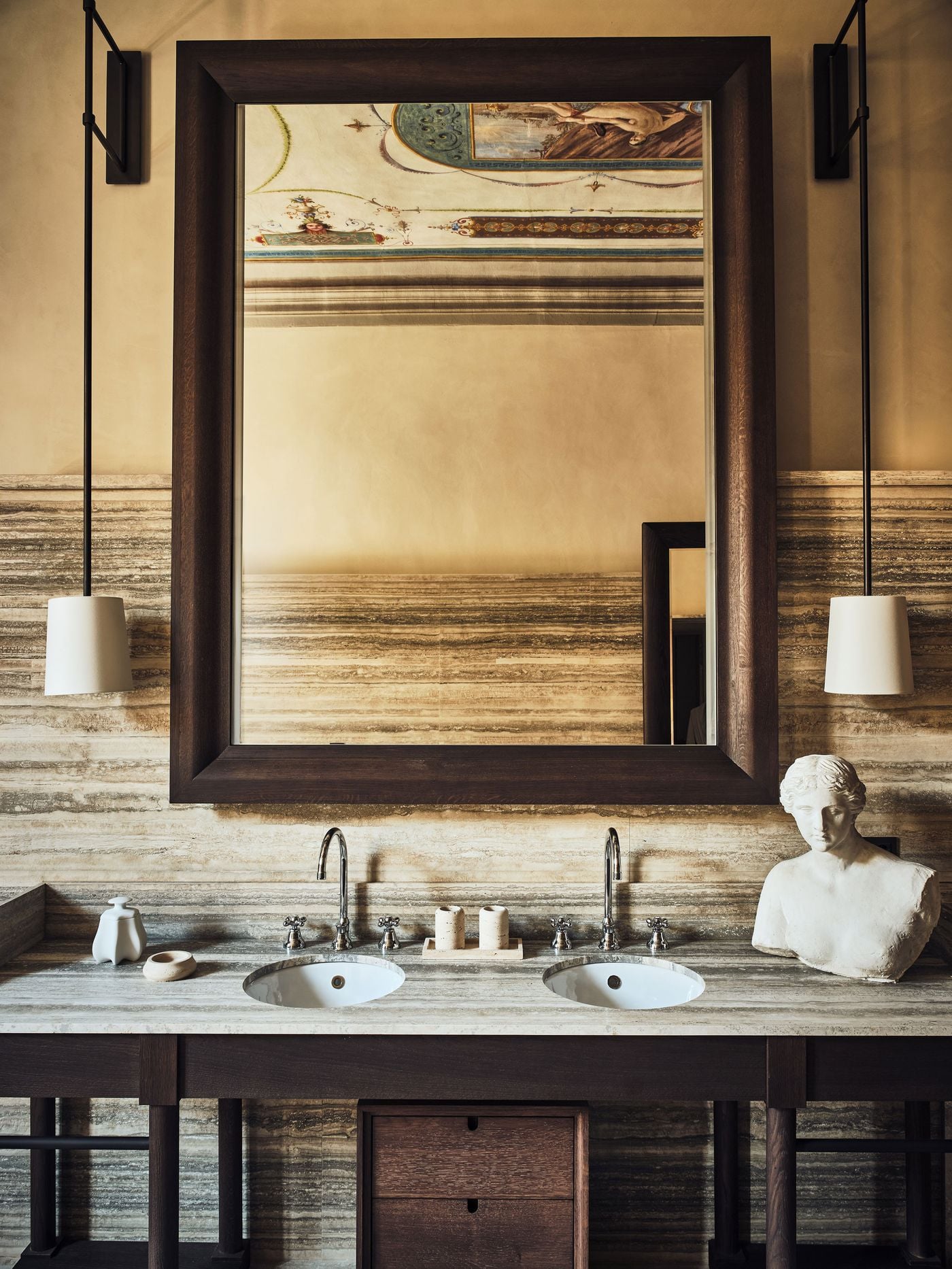 Banded travertine wraps a double vanity at La Réserve Firenze, its dark oak frame and polished nickel taps lit by a pair of suspended plaster-shaded pendants. A heavy timber mirror catches fragments of the painted ceiling above, while a classical plaster bust lends the composition a quietly sculptural presence. Banded travertine wraps a double vanity at La Réserve Firenze, its dark oak frame and polished nickel taps lit by a pair of suspended plaster-shaded pendants. A heavy timber mirror catches fragments of the painted ceiling above, while a classical plaster bust lends the composition a quietly sculptural presence.