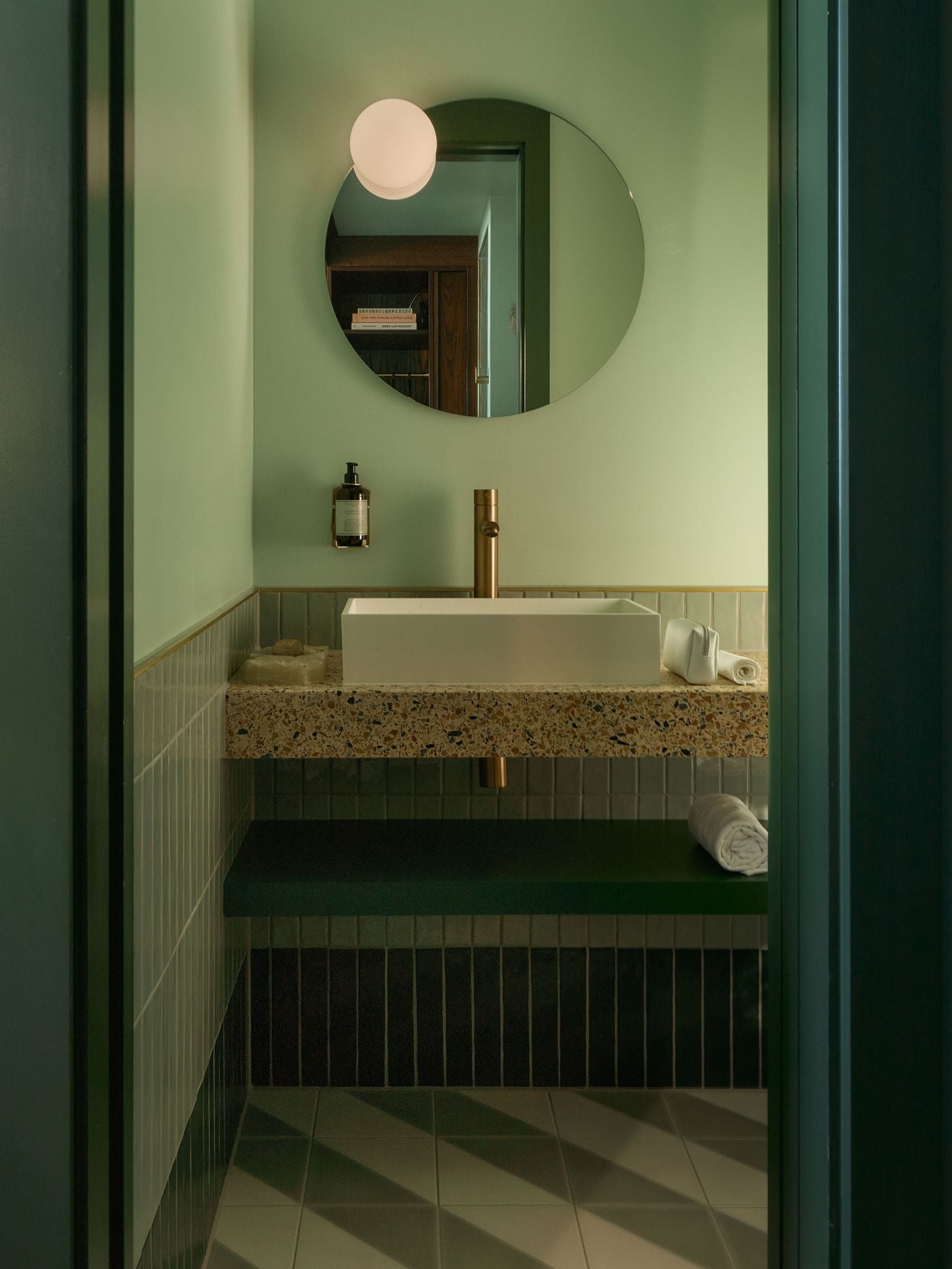 The guest-room bathroom washbasin distils the hotel’s mid-century palette into sage green, speckled terrazzo and warm brass. A circular mirror and globe light soften the compact geometry, while vertically laid tiles and a dark green shelf give the bathroom a restrained, tactile character. The guest-room bathroom washbasin distils the hotel’s mid-century palette into sage green, speckled terrazzo and warm brass. A circular mirror and globe light soften the compact geometry, while vertically laid tiles and a dark green shelf give the bathroom a restrained, tactile character.