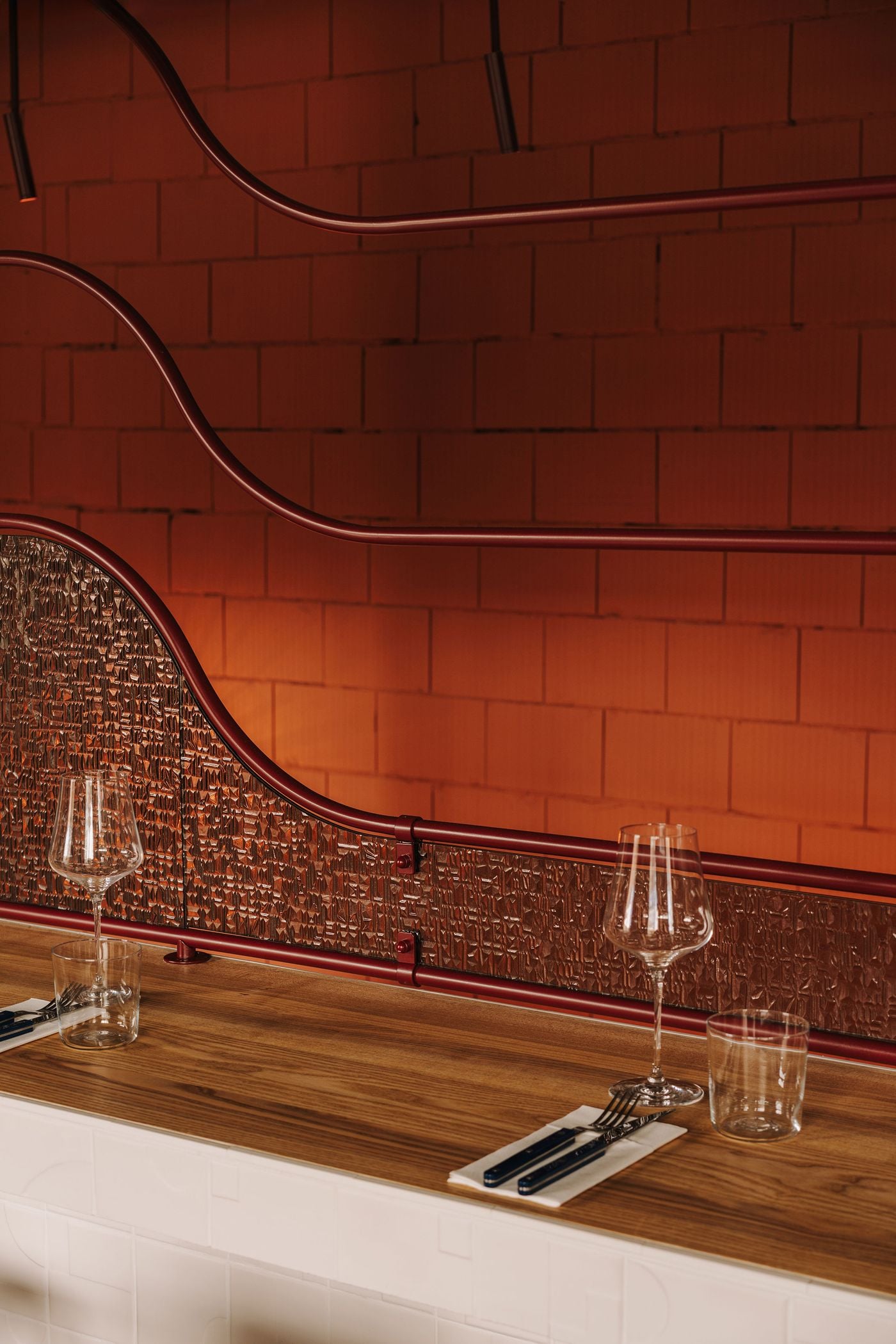 A close-up of WIN wine bar’s upstairs dining counter highlights the interplay of warm timber, white relief-patterned ceramic tiles and undulating burgundy metalwork. Textured glass panels soften the terracotta masonry backdrop, while neatly arranged wine glasses and tableware emphasise the counter’s dual function as an architectural feature and intimate dining space. A close-up of WIN wine bar’s upstairs dining counter highlights the interplay of warm timber, white relief-patterned ceramic tiles and undulating burgundy metalwork. Textured glass panels soften the terracotta masonry backdrop, while neatly arranged wine glasses and tableware emphasise the counter’s dual function as an architectural feature and intimate dining space.
