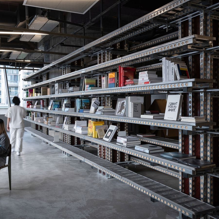 An expansive view of the 889 GLO Art Space showcases a lengthy, multi-tiered shelving unit crafted from industrial metal components, filled with a diverse collection of books and art objects. A person walks along the concrete floor, highlighting the impressive scale and the raw, yet curated, brutalist-industrial design of the space. An expansive view of the 889 GLO Art Space showcases a lengthy, multi-tiered shelving unit crafted from industrial metal components, filled with a diverse collection of books and art objects. A person walks along the concrete floor, highlighting the impressive scale and the raw, yet curated, brutalist-industrial design of the space.
