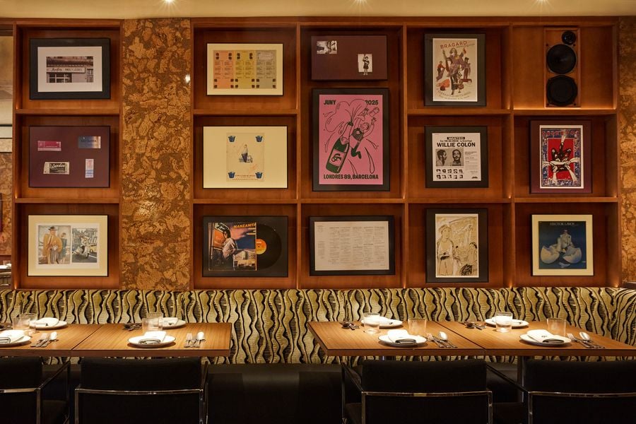 A well-composed wide shot of a dining space showcasing a striking gallery wall. The dark wooden shelving unit contains an assortment of framed posters, records, and photographs. The lively patterned seating below and simple wooden tables with black chairs complete the cohesive and visually rich aesthetic.