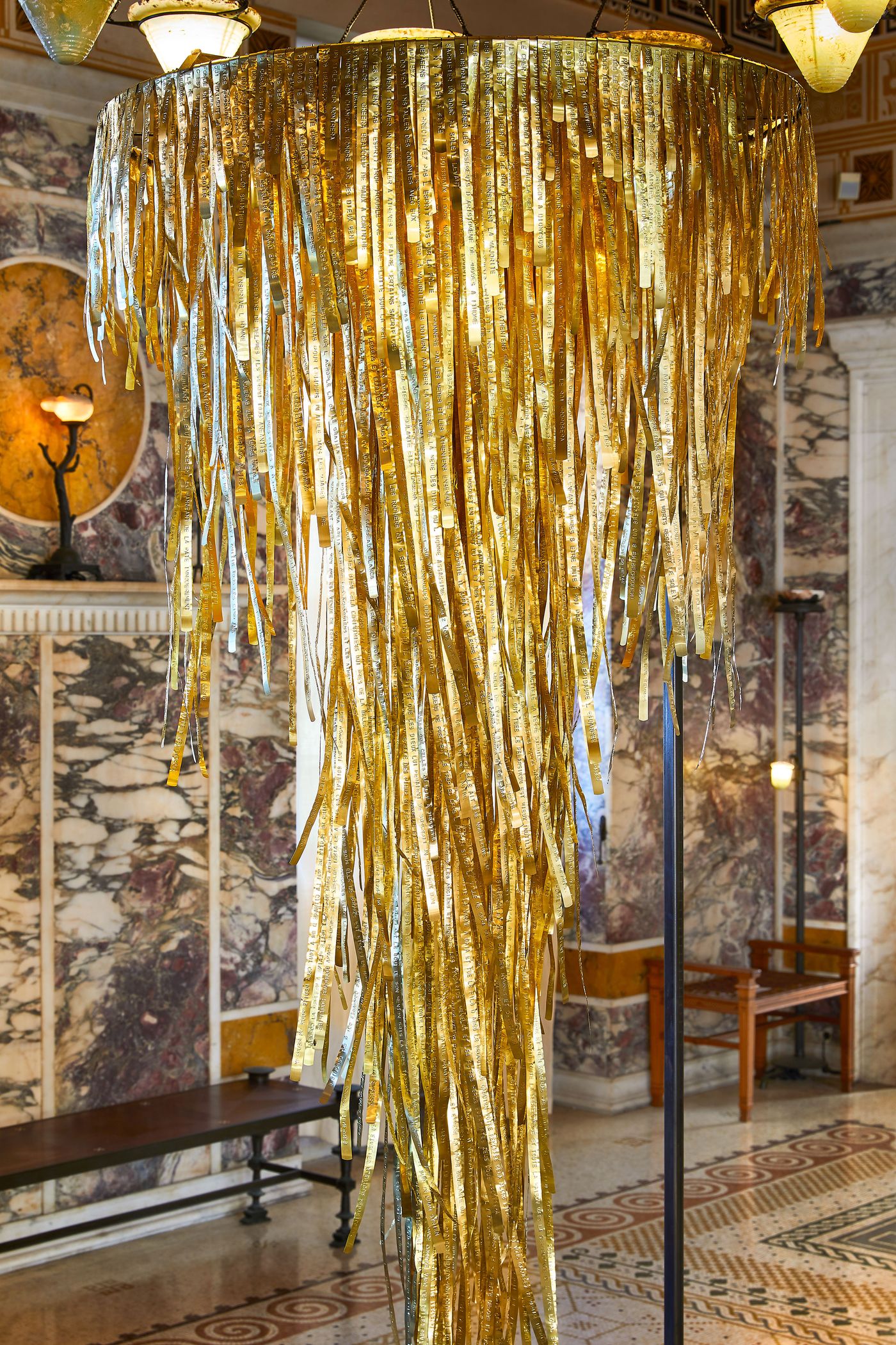 A close-up of the cascading golden sculpture by Gabriel Leger. The artwork, a dense flow of shimmering, text-etched strips, hangs from a chandelier, creating a waterfall-like effect. The piece is a central figure in a hall with striking red-veined marble and intricate wall sconces, showcasing a fusion of classical and contemporary design.