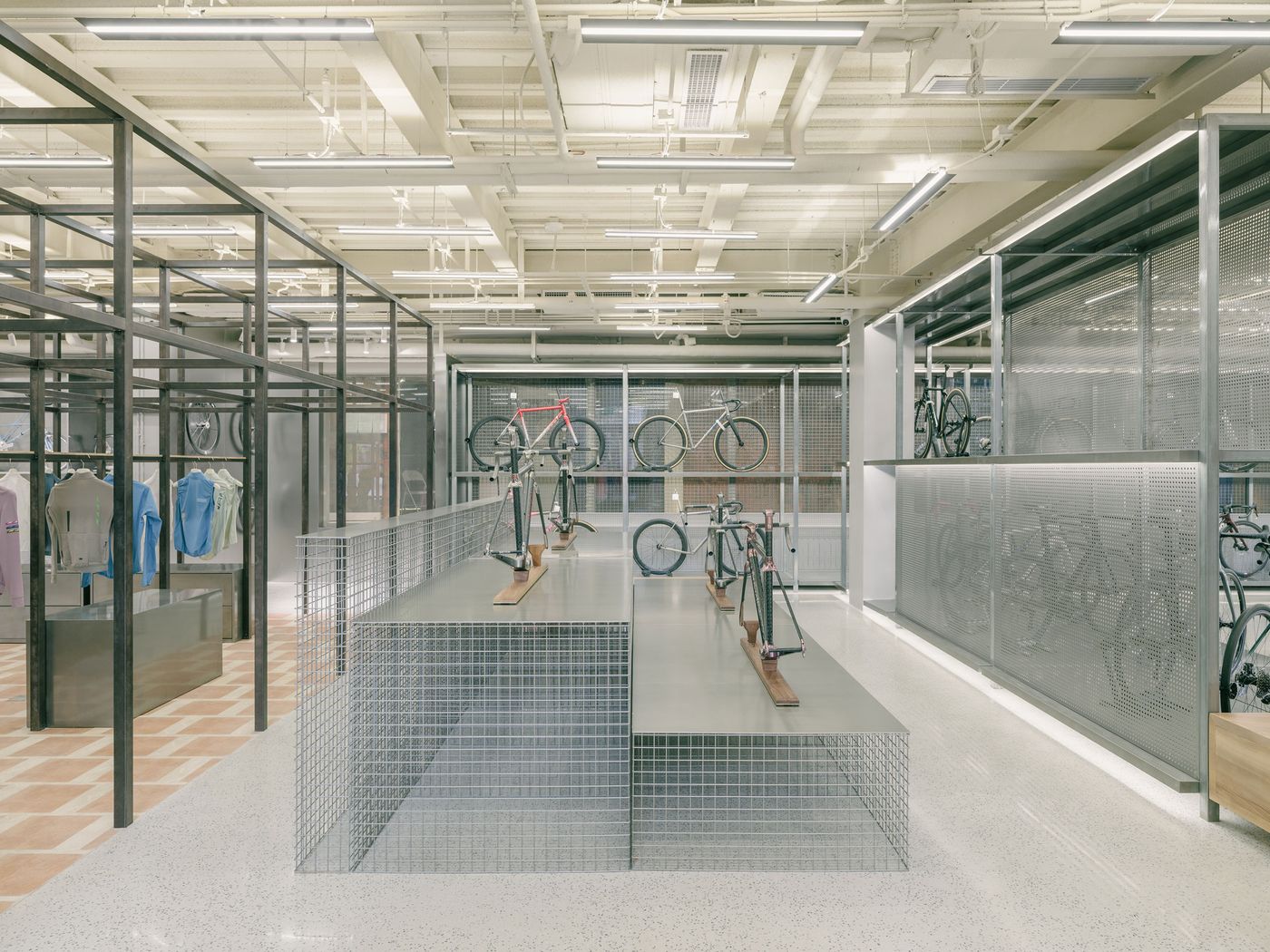 A vast and coherent retail space showcases a blend of industrial and minimalist design. Sleek, low-profile wire mesh platforms display bicycles and gear, while a grid of black metal framing defines areas for apparel. The polished concrete floor with terracotta tile inlays contrasts with the exposed white ceiling infrastructure, creating a contemporary, functional aesthetic. A vast and coherent retail space showcases a blend of industrial and minimalist design. Sleek, low-profile wire mesh platforms display bicycles and gear, while a grid of black metal framing defines areas for apparel. The polished concrete floor with terracotta tile inlays contrasts with the exposed white ceiling infrastructure, creating a contemporary, functional aesthetic.