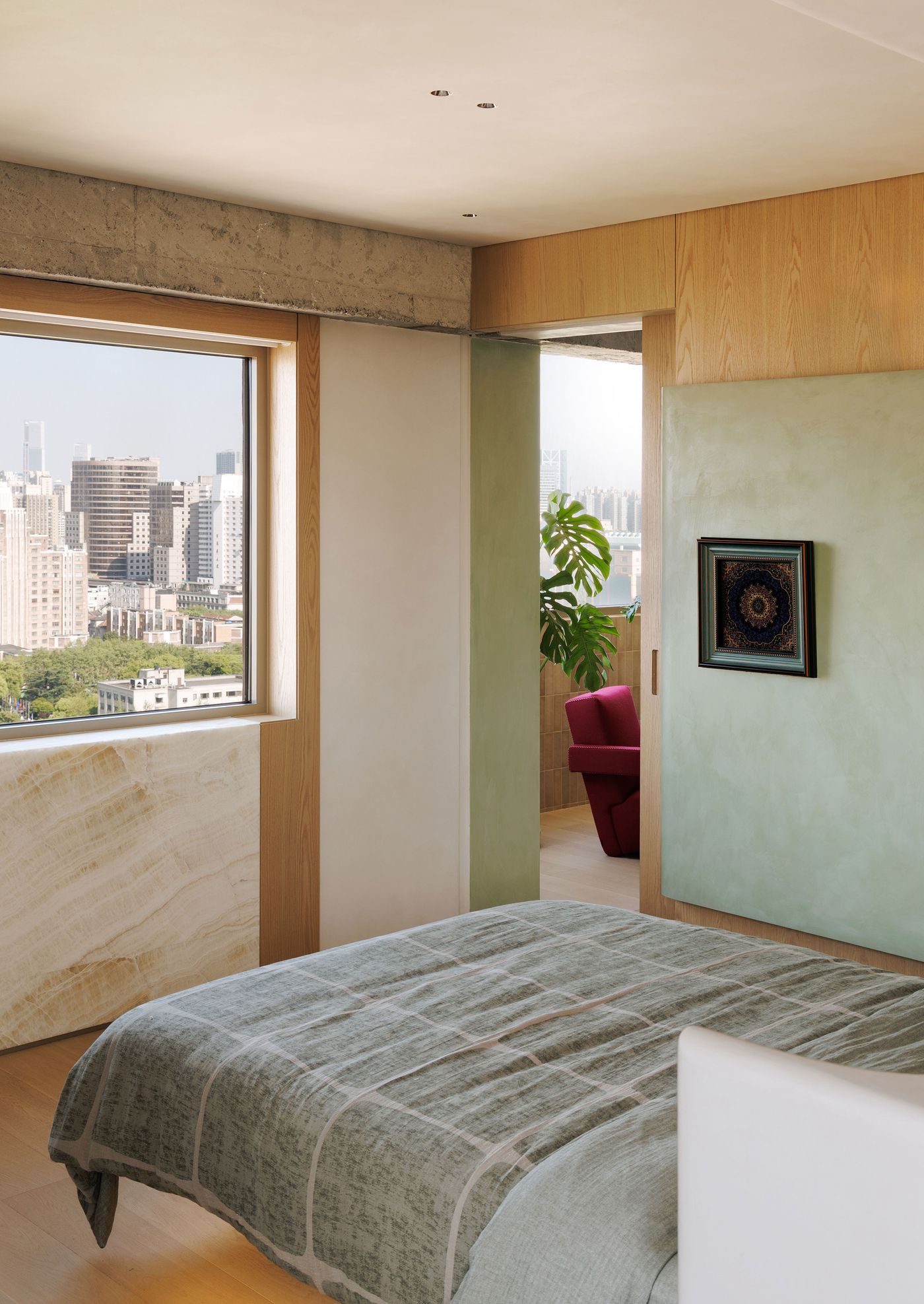 A minimalist bedroom designed for tranquility and urban connection. A soft green wall panel with framed art adds a subtle color accent against the warm wood and exposed concrete beam. Expansive windows reveal the Shanghai skyline, while a luxurious marble panel below the window introduces natural texture. A vibrant red armchair is visible in the adjacent room, inviting relaxation. A minimalist bedroom designed for tranquility and urban connection. A soft green wall panel with framed art adds a subtle color accent against the warm wood and exposed concrete beam. Expansive windows reveal the Shanghai skyline, while a luxurious marble panel below the window introduces natural texture. A vibrant red armchair is visible in the adjacent room, inviting relaxation.