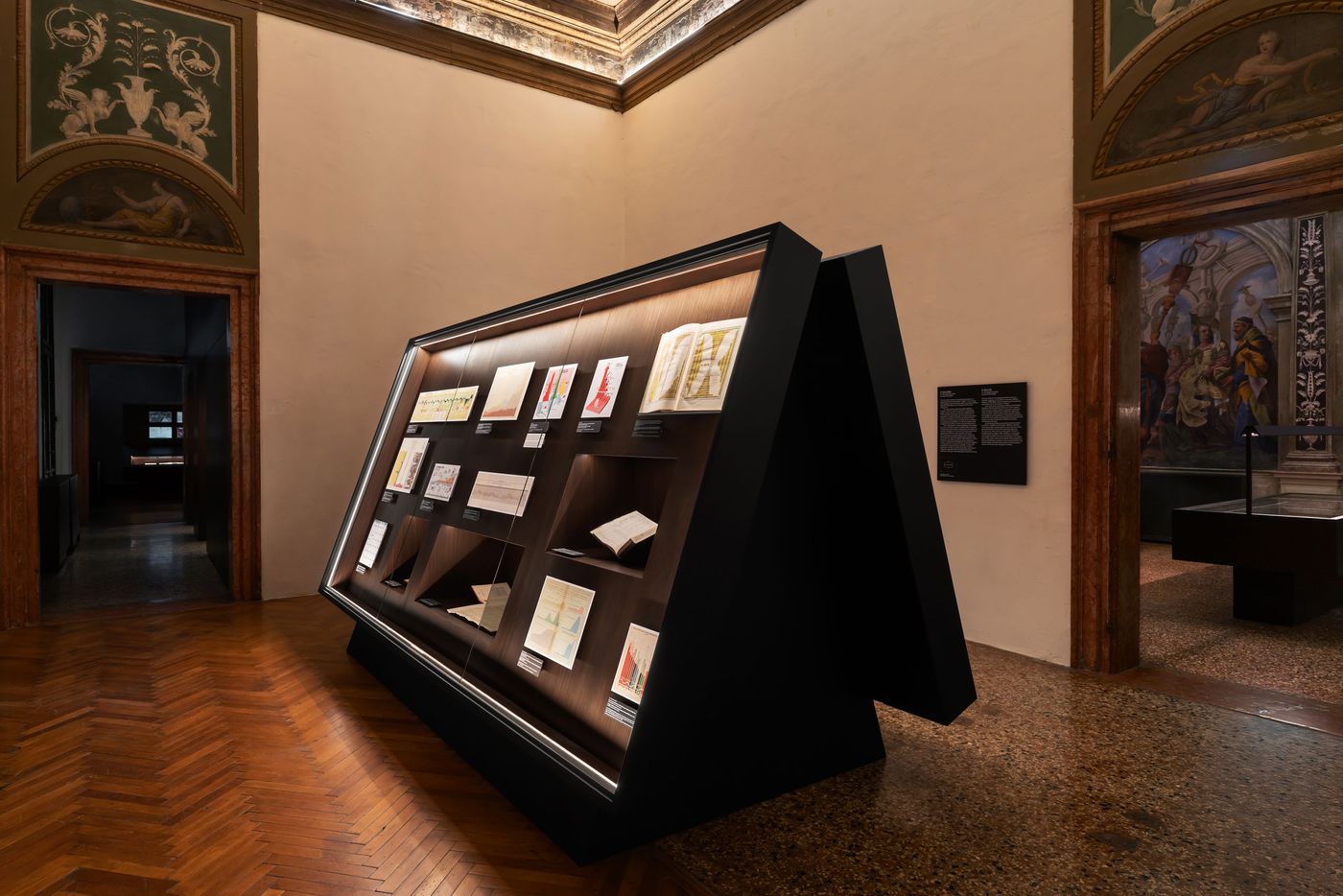 A modern display case showcases various colorful art sketches in a museum setting with an ornate ceiling and wooden flooring. A modern display case showcases various colorful art sketches in a museum setting with an ornate ceiling and wooden flooring.