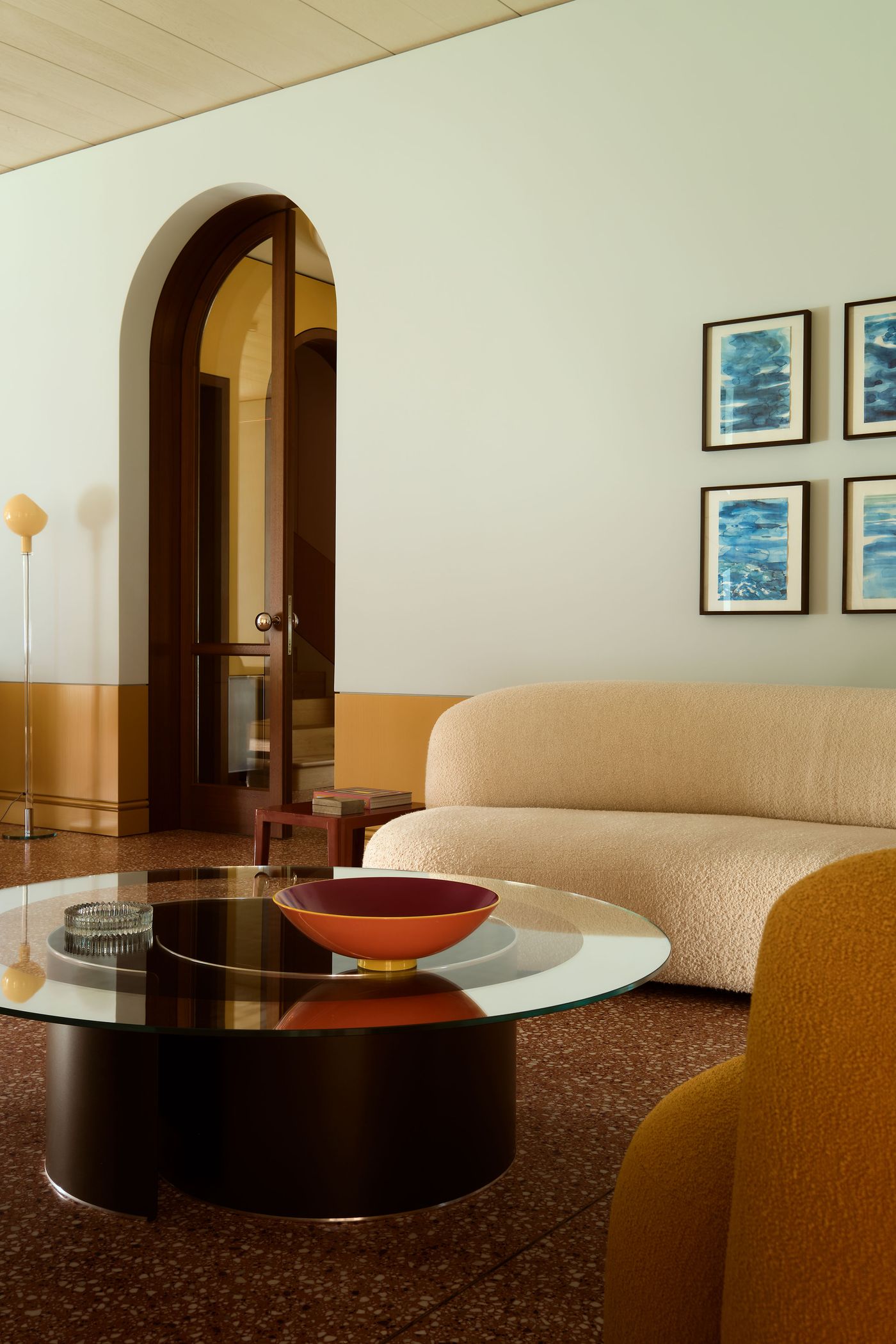 A modern living room featuring a beige sofa, circular glass coffee table with a red bowl, and artistic wall decor, with an arched doorway.