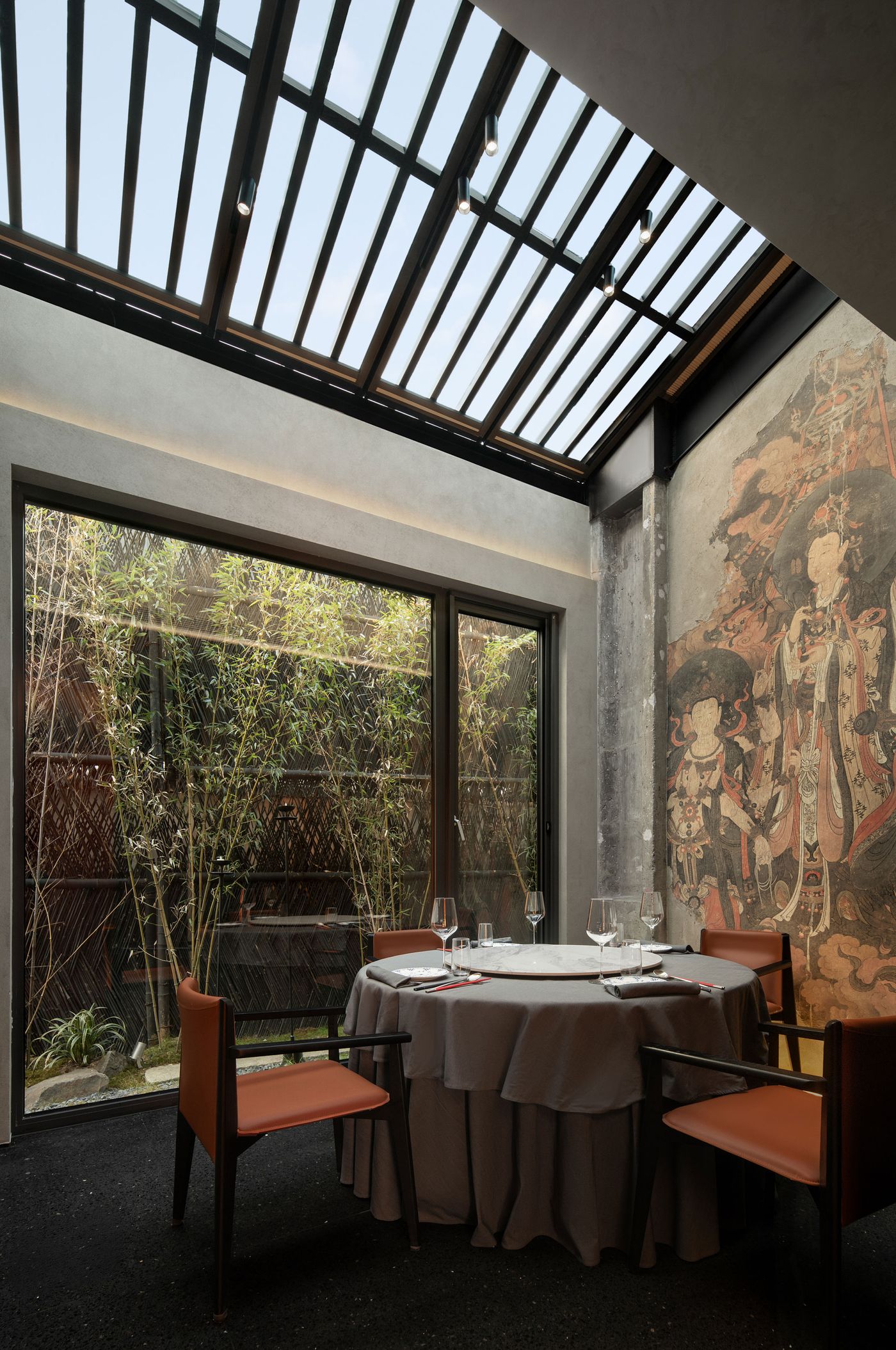 Cozy dining area featuring a round table with elegant tableware, surrounded by a wall mural and natural light from a slatted roof. Cozy dining area featuring a round table with elegant tableware, surrounded by a wall mural and natural light from a slatted roof.