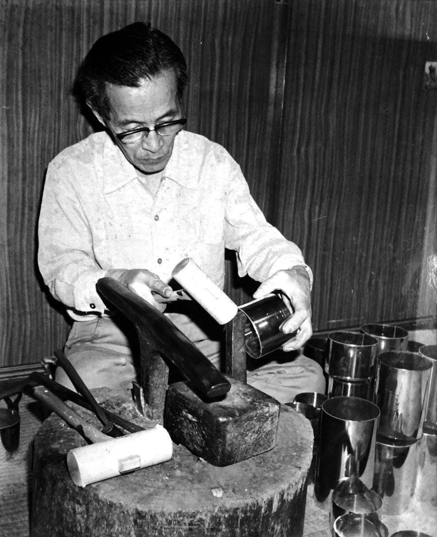 An artisan works at a wooden stump with various metal cups and tools surrounding him, showcasing traditional craftsmanship. An artisan works at a wooden stump with various metal cups and tools surrounding him, showcasing traditional craftsmanship.