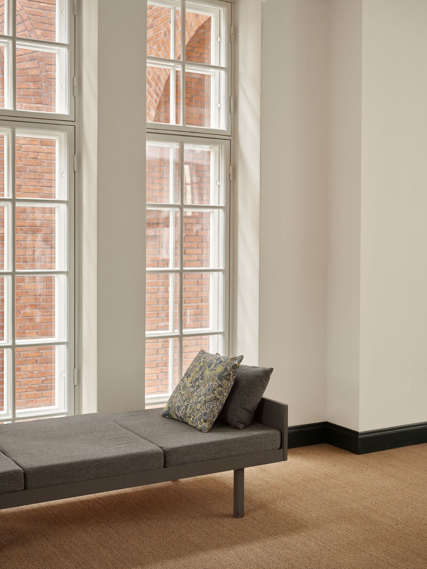 A minimalist grey daybed with a patterned cushion is positioned in a sunlit corner next to tall, multi-pane windows that reveal a brick building exterior. The stark white walls and natural flooring create a serene and understated resting space.