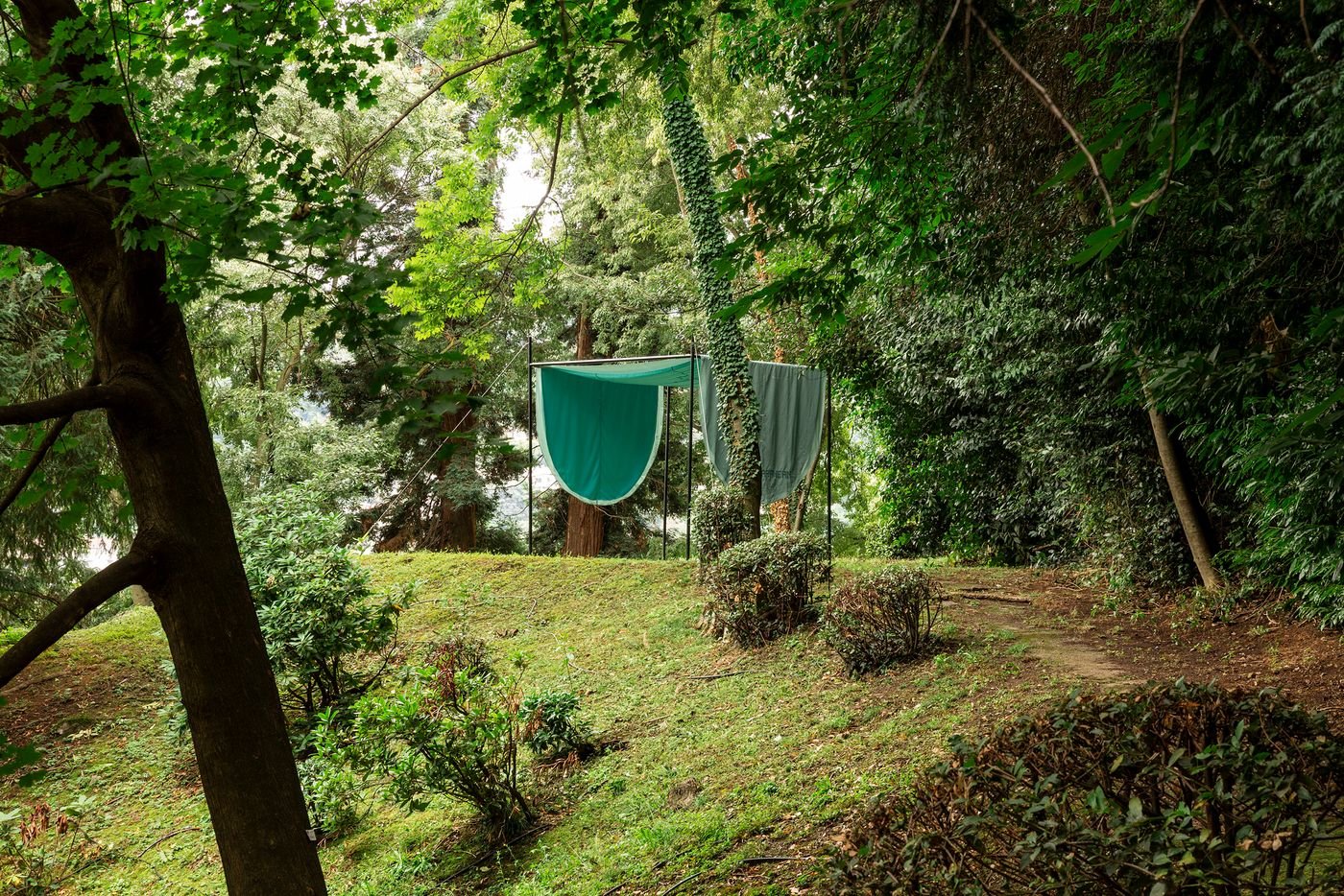 A tranquil forest clearing reveals a four-post structure covered in emerald green fabric, draped to create soft, organic shapes. This installation is set against a backdrop of dense foliage and tall trees, with a winding path leading through the scene. The interplay of natural light, lush greenery, and the flowing fabric creates a serene and almost ethereal atmosphere, evoking a sense of natural artistry.