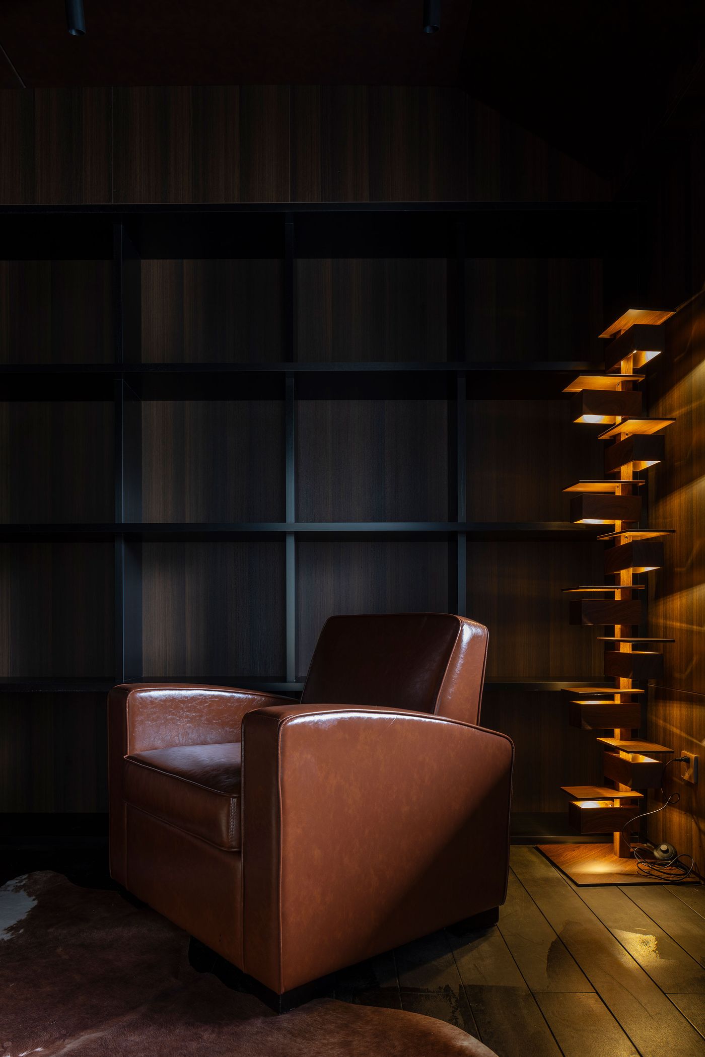 A warm, inviting interior nook within Wutopia Lab's Lake House showcases a rich brown leather armchair set against a dark wood paneled wall and integrated shelving. A distinctive, illuminated vertical sculpture on the right, crafted from staggered golden elements, casts an ambient glow, creating a sophisticated and contemplative atmosphere that blends modern design with a subtle sense of luxury.