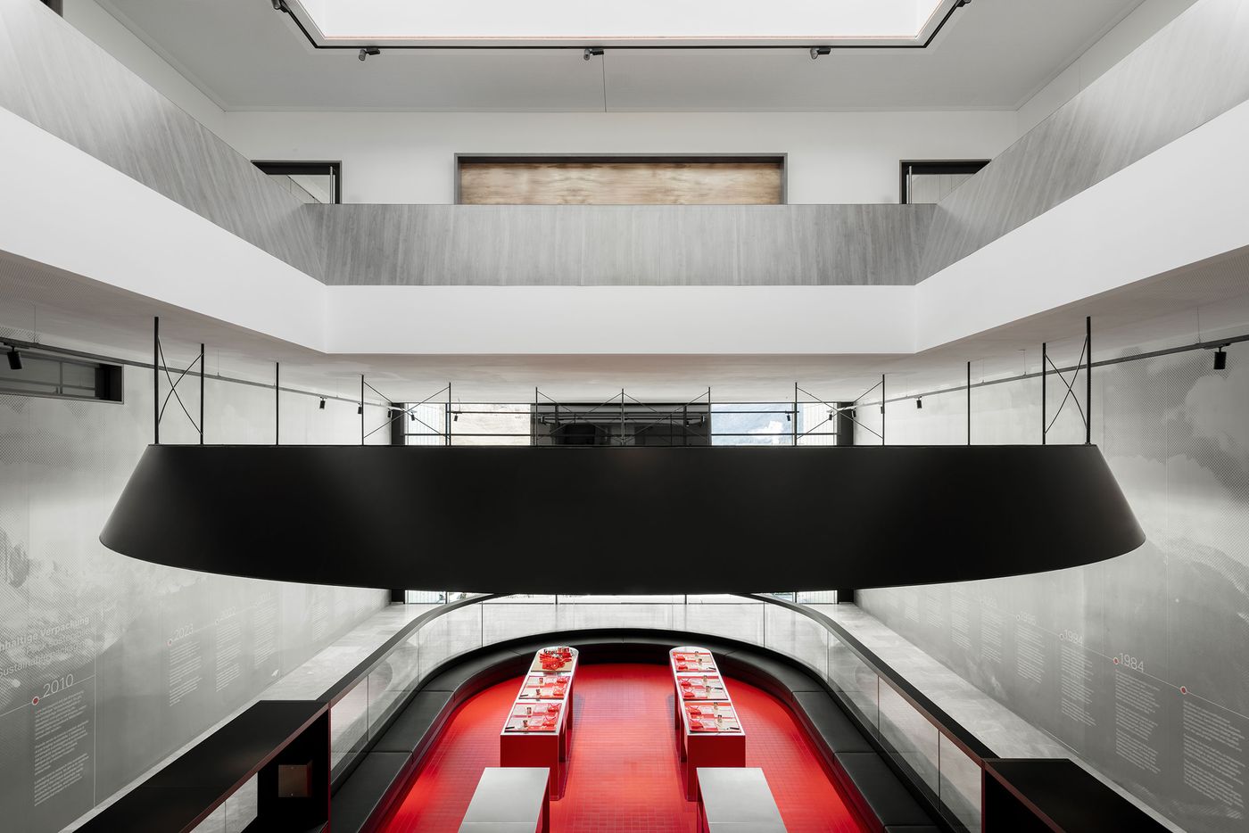 A monumental double-height space features floating black ceiling elements above curved exhibition walls displaying historical timeline. The dramatic scale and sophisticated material palette create museum-quality environment where corporate history becomes cultural narrative through precise architectural staging and lighting design. A monumental double-height space features floating black ceiling elements above curved exhibition walls displaying historical timeline. The dramatic scale and sophisticated material palette create museum-quality environment where corporate history becomes cultural narrative through precise architectural staging and lighting design.
