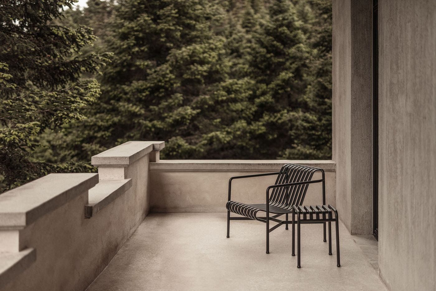 A minimalist outdoor balcony space extends from the hotel, featuring a sleek, black metal slatted chair and a matching small side table. The robust, light-colored concrete balustrade and wall contrast with the dense, dark green pine forest in the background, creating a tranquil setting for contemplation and connection with nature. A minimalist outdoor balcony space extends from the hotel, featuring a sleek, black metal slatted chair and a matching small side table. The robust, light-colored concrete balustrade and wall contrast with the dense, dark green pine forest in the background, creating a tranquil setting for contemplation and connection with nature.