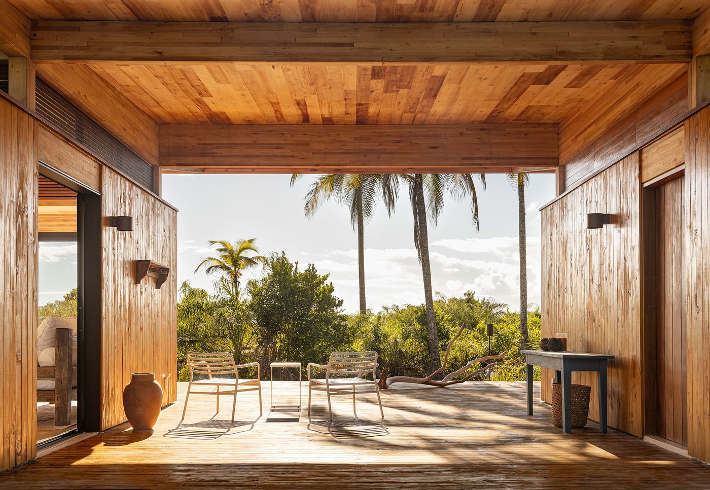 A sheltered outdoor lounge area features an expansive wooden deck flanked by warm timber walls, opening directly to a sun-drenched tropical landscape. Two woven chairs and a small table invite relaxation, embodying a harmonious connection to the surrounding nature. A sheltered outdoor lounge area features an expansive wooden deck flanked by warm timber walls, opening directly to a sun-drenched tropical landscape. Two woven chairs and a small table invite relaxation, embodying a harmonious connection to the surrounding nature.