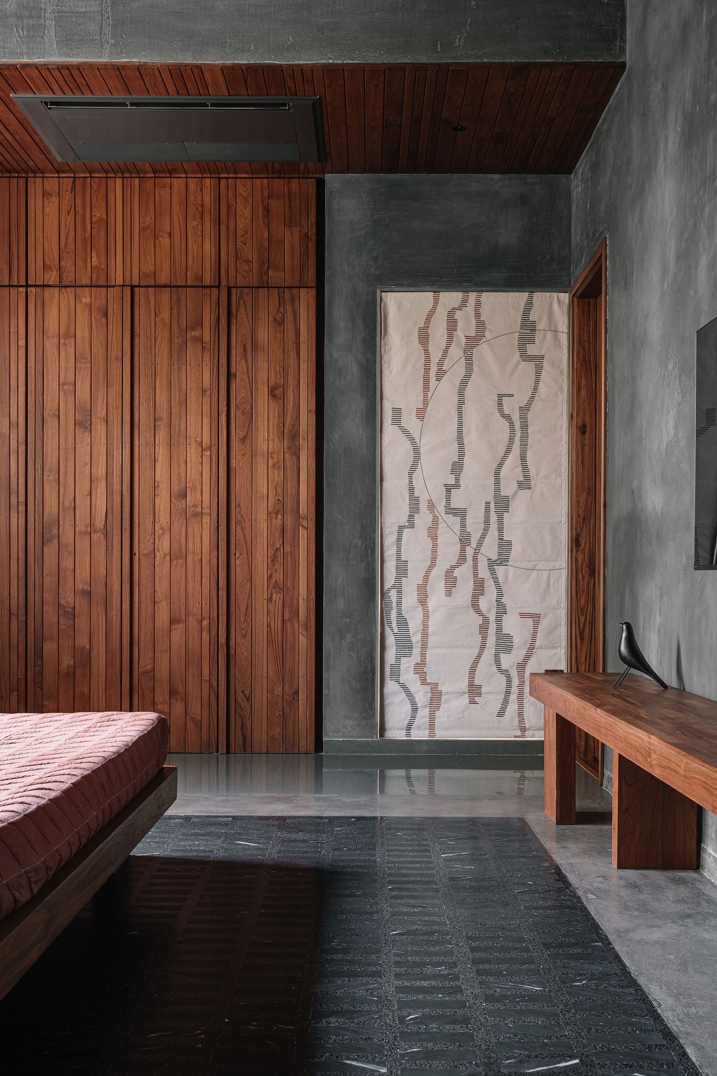 A sophisticated bedroom with raw concrete walls and rich wooden built-in wardrobes, offering a contrast of textures. A unique abstract artwork on a textile panel adorns one wall. The low-profile bed and a long wooden bench maintain a minimalist aesthetic, while the dark floor anchors the earthy and elegant color palette.