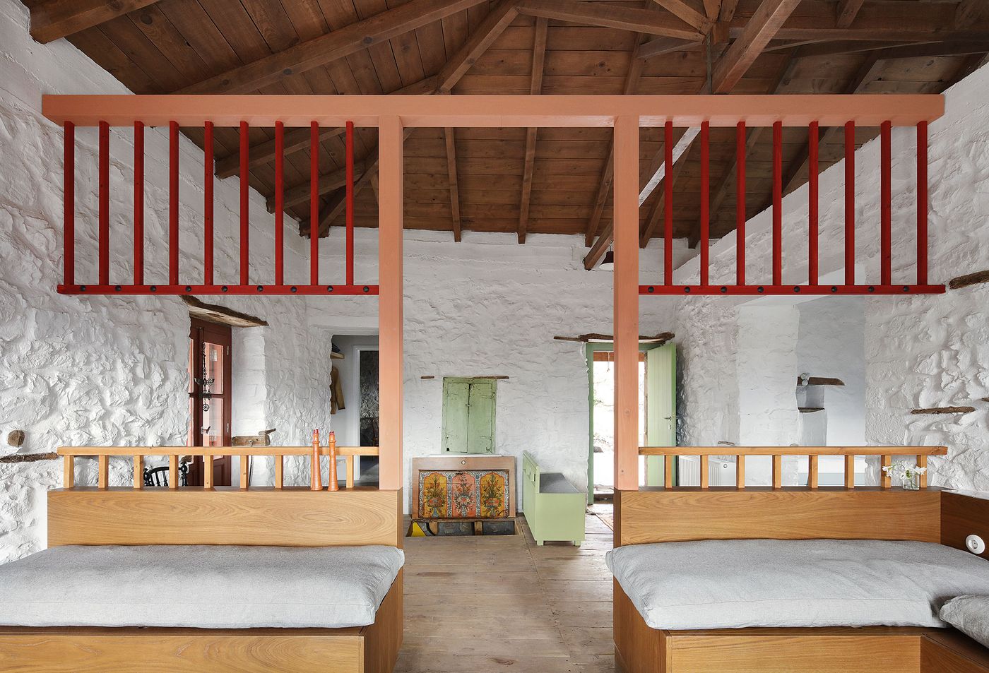 Weathered mint-green traditional shutters provide authentic Mediterranean color against the whitewashed stone walls, while coral-red contemporary elements establish visual continuity. The dormitory-style arrangement celebrates communal living through carefully orchestrated furniture placement and mezzanine relationships.
