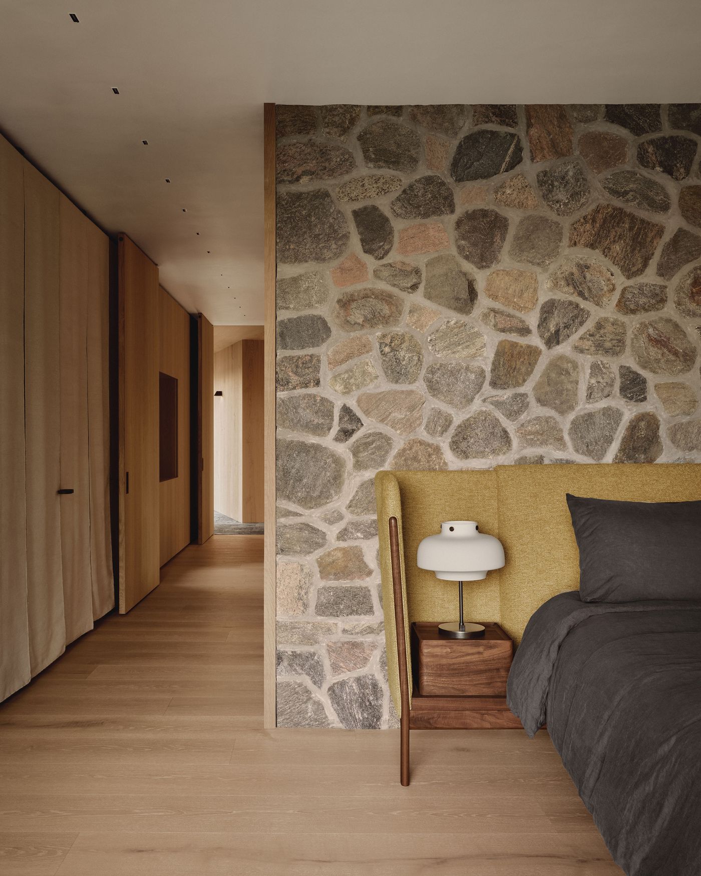 Cozy bedroom featuring a stone wall, a soft yellow headboard, dark bedding, and a wooden nightstand with a modern lamp. Cozy bedroom featuring a stone wall, a soft yellow headboard, dark bedding, and a wooden nightstand with a modern lamp.