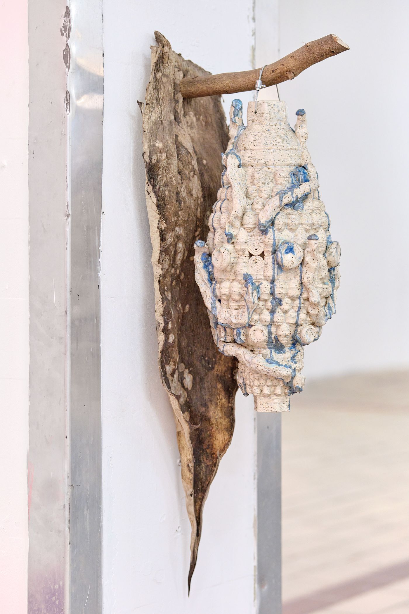 A textured sculpture resembling a hanging lantern, adorned with blue accents, mounted on a piece of wood, displayed in a minimalist space.