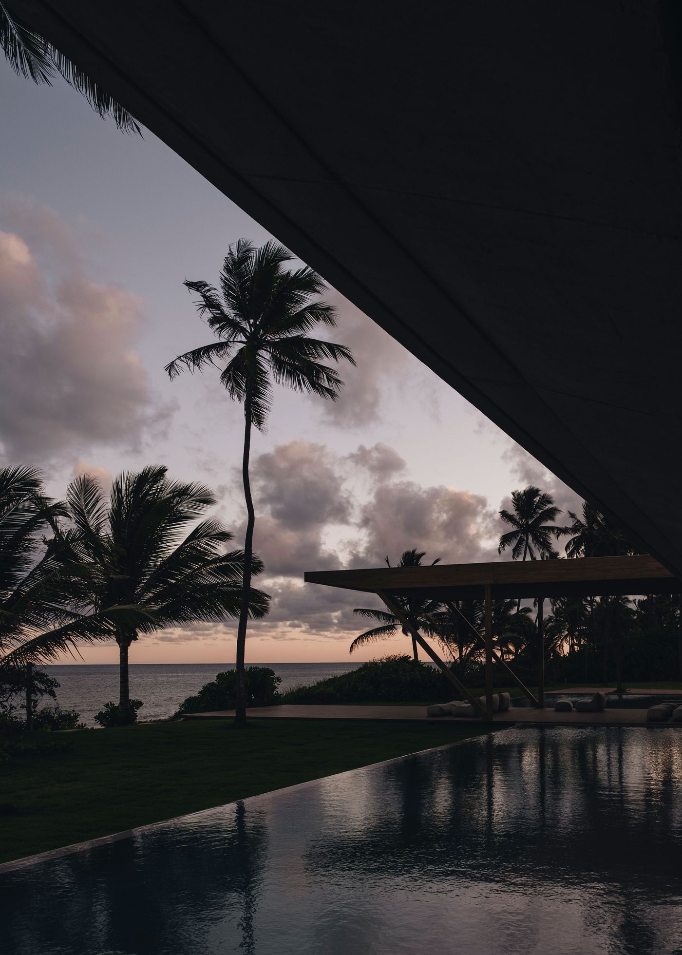 A tranquil coastal scene at dusk, featuring palm trees, a serene pool reflecting the sky, and clouds scattered above the ocean horizon.