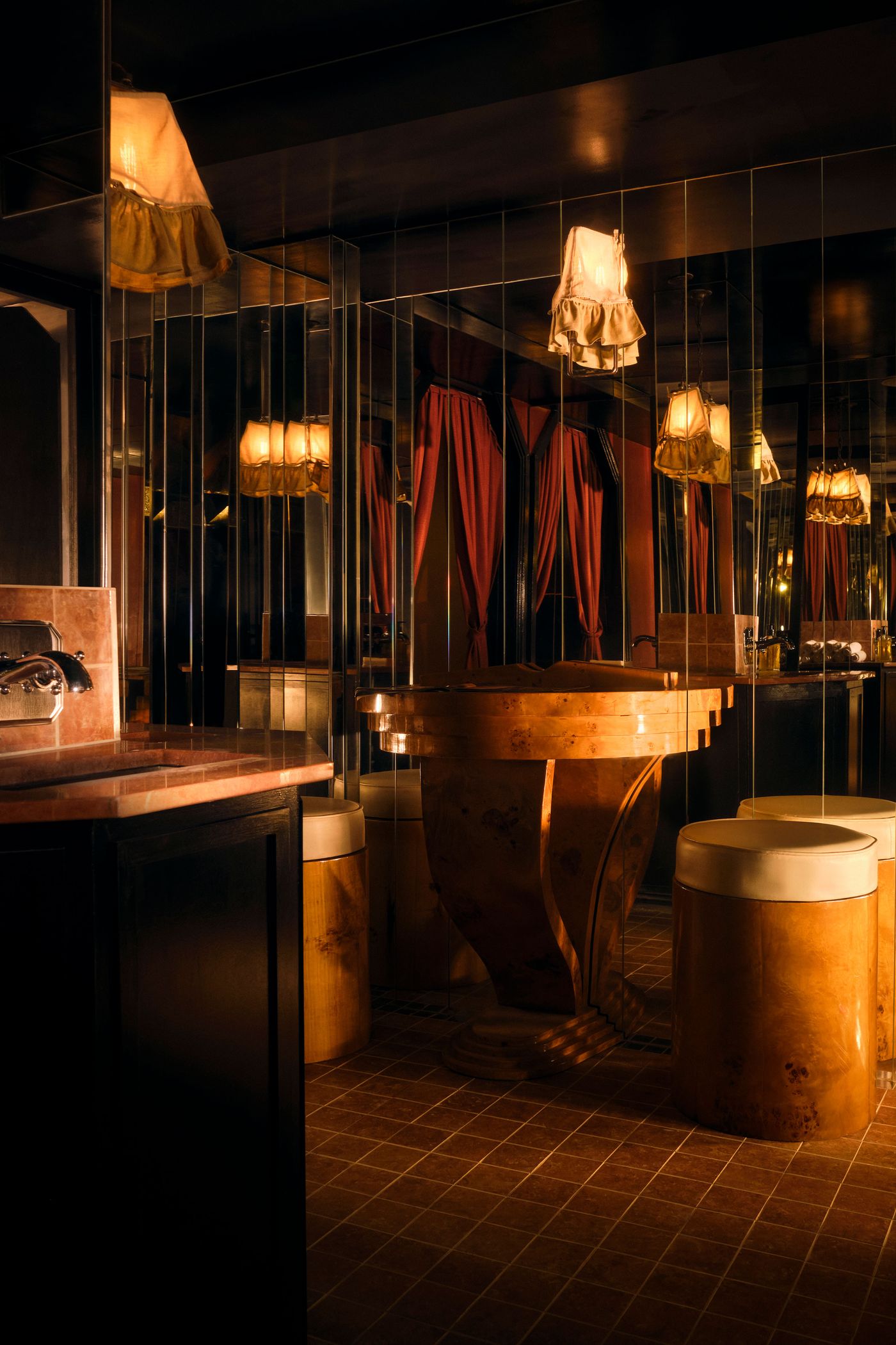The opulent and reflective washroom of The Roses of Elagabalus, featuring a central art-deco style wooden vanity with curvilinear forms. Mirrored walls multiply the warm glow from vintage pendant lamps with ruffled shades and create a sense of expansive luxury. Pink marble countertops and a checkered floor complete the decadent, vintage-retro aesthetic.