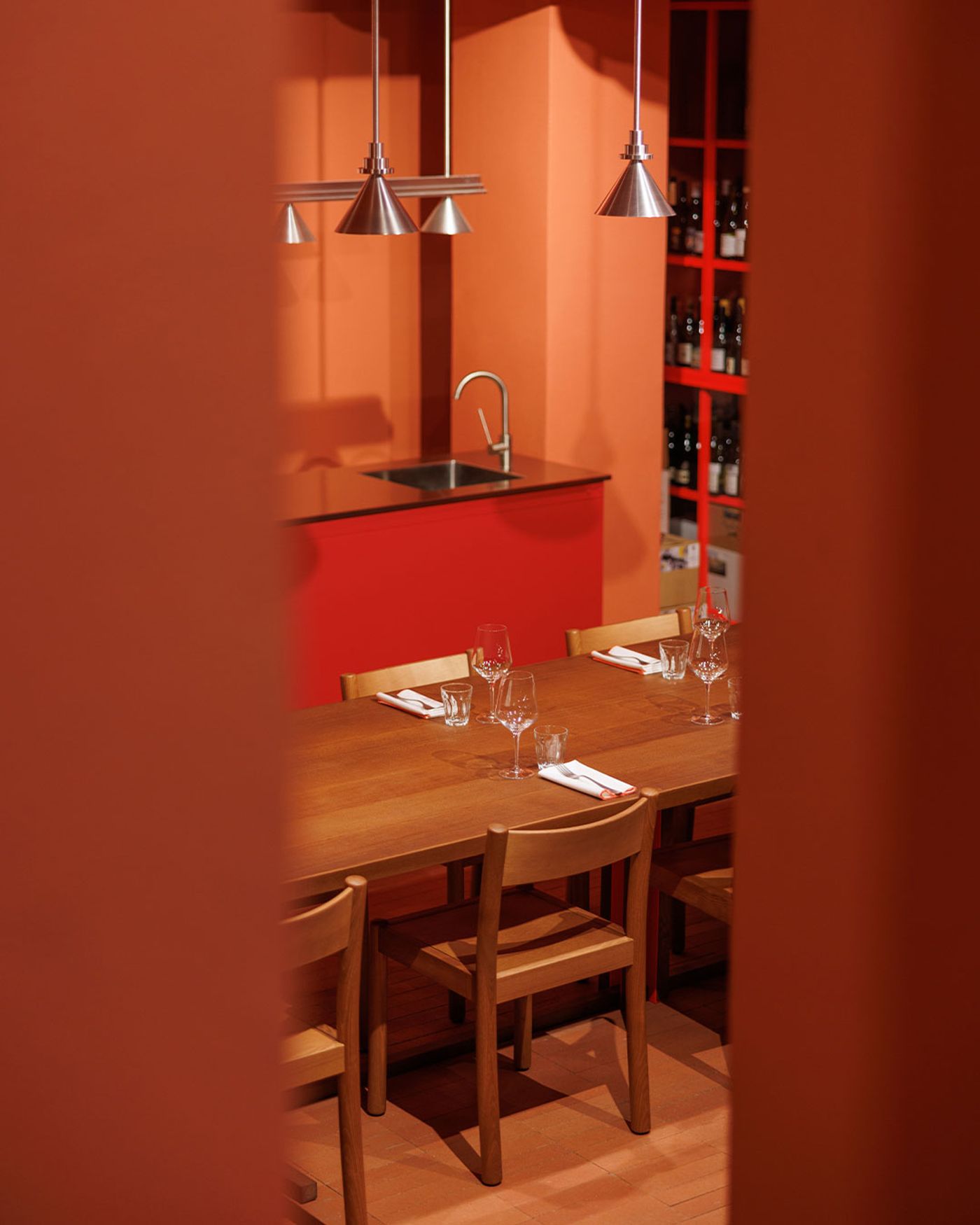 A captivating view of Santo Palato's dining area by Naessi Studio, framed by a soft, warm red foreground element. A long, polished wooden communal table is meticulously set with glassware and crisp white napkins. In the background, a striking red wine rack filled with bottles and a minimalist counter with a sink are visible, all bathed in the warm, inviting glow of industrial pendant lights, reflecting a modern Roman trattoria aesthetic. A captivating view of Santo Palato's dining area by Naessi Studio, framed by a soft, warm red foreground element. A long, polished wooden communal table is meticulously set with glassware and crisp white napkins. In the background, a striking red wine rack filled with bottles and a minimalist counter with a sink are visible, all bathed in the warm, inviting glow of industrial pendant lights, reflecting a modern Roman trattoria aesthetic.