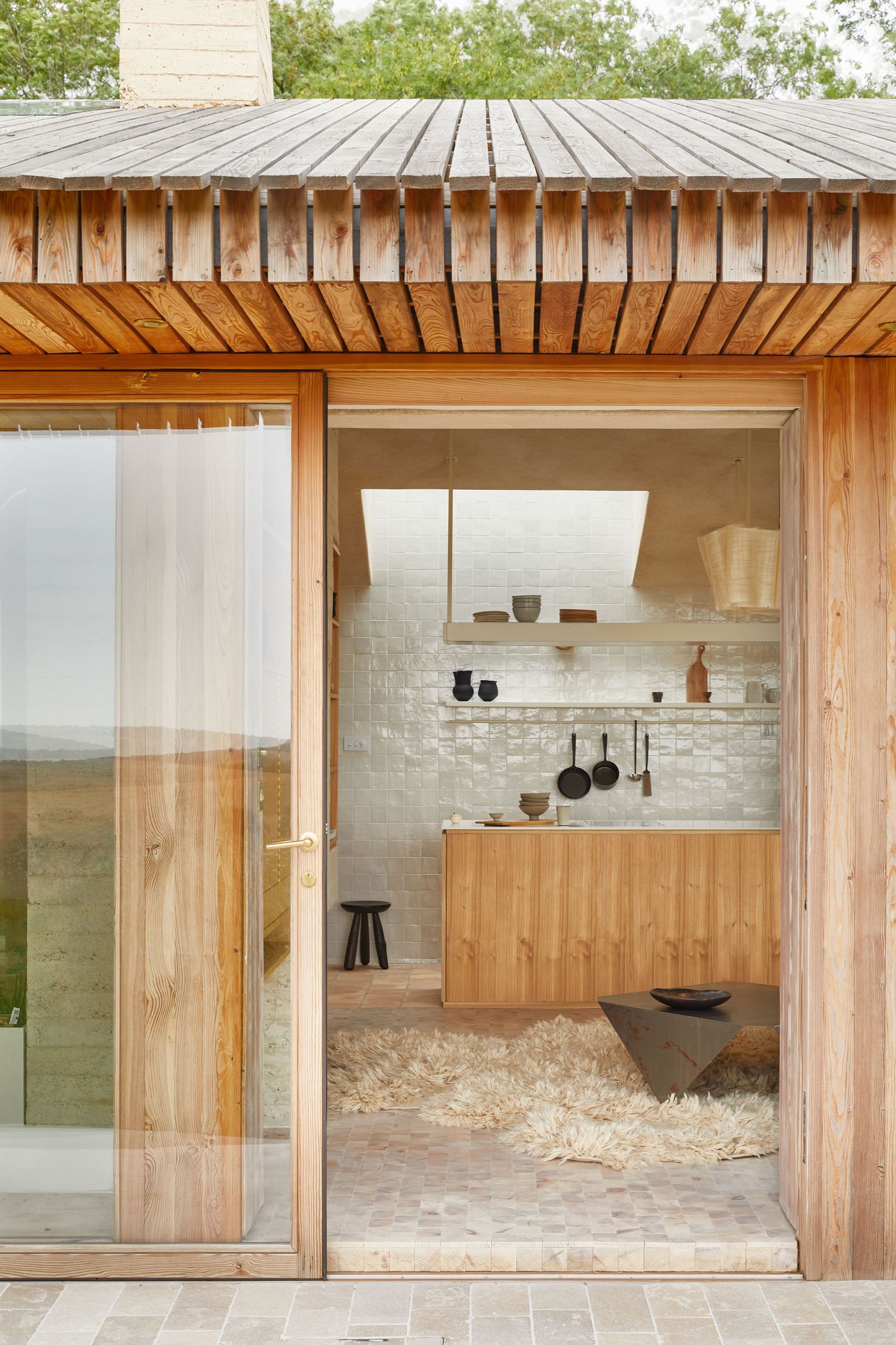 Looking into The Maker's Barn by Hutch Design from outside, a large timber-framed sliding glass door reveals a contemporary kitchen. The interior features light wooden cabinetry, a tiled backsplash, and open shelving, illuminated by a large skylight. A textured, shaggy rug anchors the space, inviting a seamless transition between indoor comfort and the natural exterior. Looking into The Maker's Barn by Hutch Design from outside, a large timber-framed sliding glass door reveals a contemporary kitchen. The interior features light wooden cabinetry, a tiled backsplash, and open shelving, illuminated by a large skylight. A textured, shaggy rug anchors the space, inviting a seamless transition between indoor comfort and the natural exterior.
