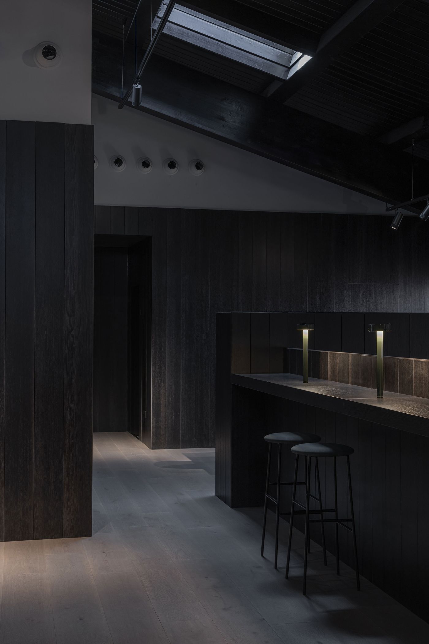 A dimly lit modern interior featuring dark wooden walls, a sleek bar with illuminated lamps, and minimalistic black stools.
