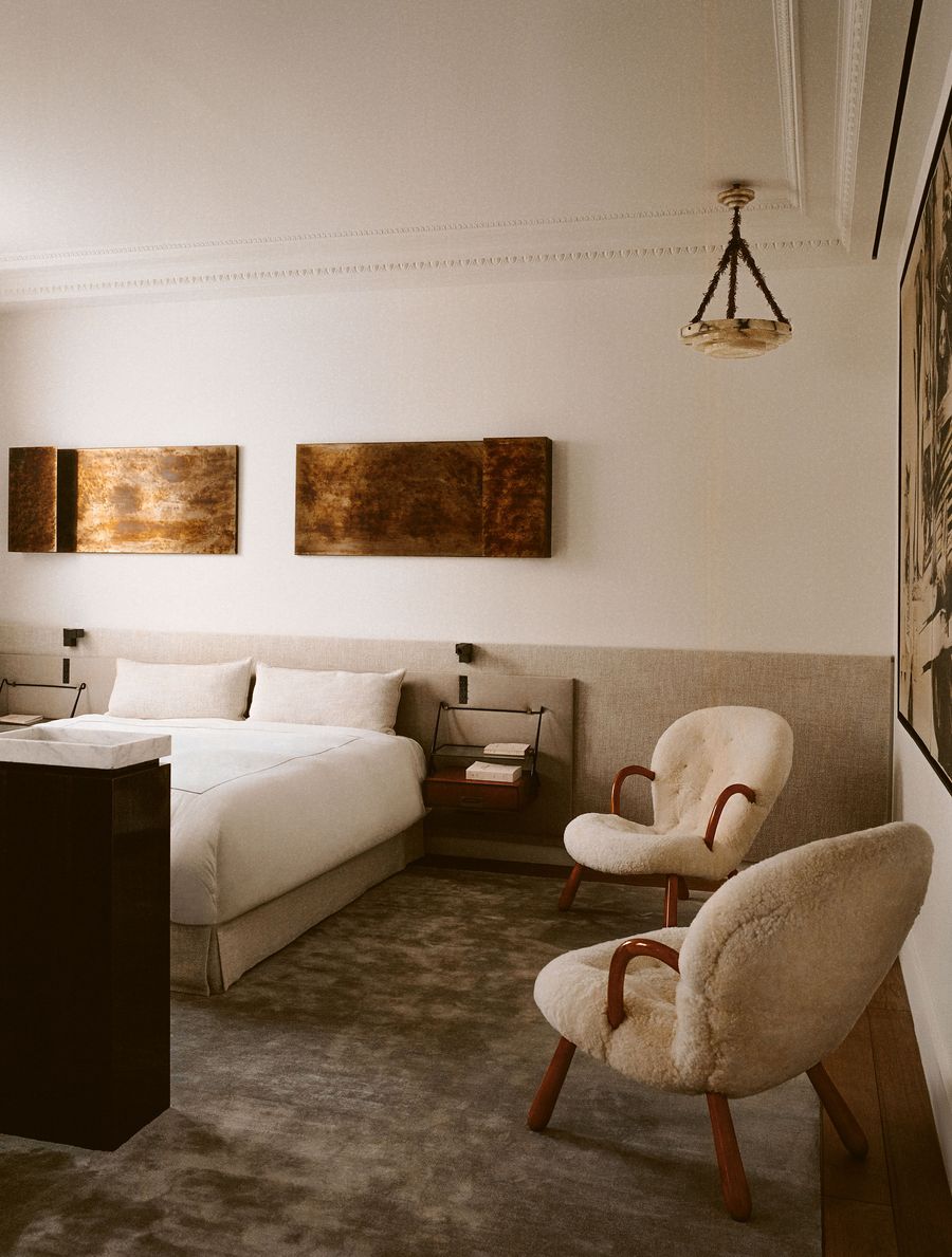 A tranquil bedroom with a large, inviting bed featuring crisp white linens and an upholstered headboard in a neutral tone. Two unique, plush sheepskin-covered armchairs with warm wooden frames add a touch of mid-century modern comfort. The walls display two abstract, textured artworks in metallic hues. A tranquil bedroom with a large, inviting bed featuring crisp white linens and an upholstered headboard in a neutral tone. Two unique, plush sheepskin-covered armchairs with warm wooden frames add a touch of mid-century modern comfort. The walls display two abstract, textured artworks in metallic hues.