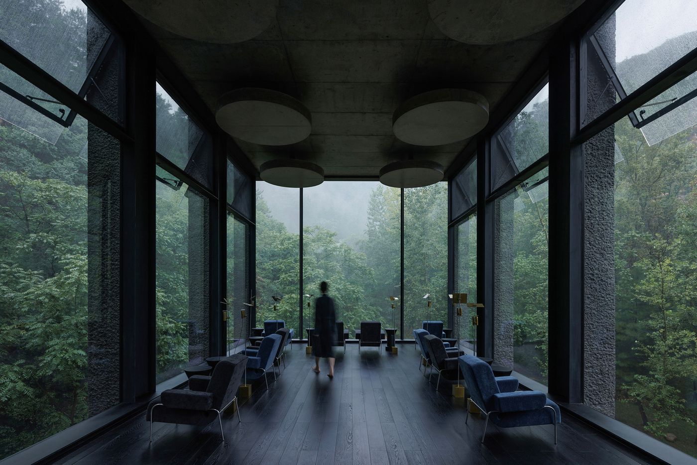 A person walks through a modern, minimalist lounge with large windows showcasing lush greenery and rain outside. A person walks through a modern, minimalist lounge with large windows showcasing lush greenery and rain outside.
