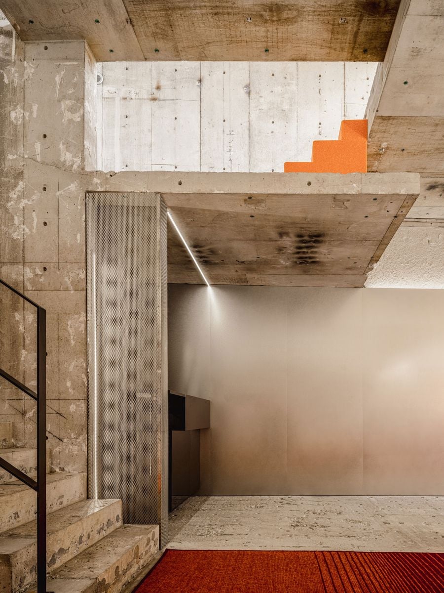 A quieter transitional area combines unfinished concrete stairs and slabs with a large expanse of smooth metallic wall cladding. A perforated metal screen conceals services beside the staircase, while a small flight of vivid orange steps appears above, injecting colour into the otherwise muted industrial palette.