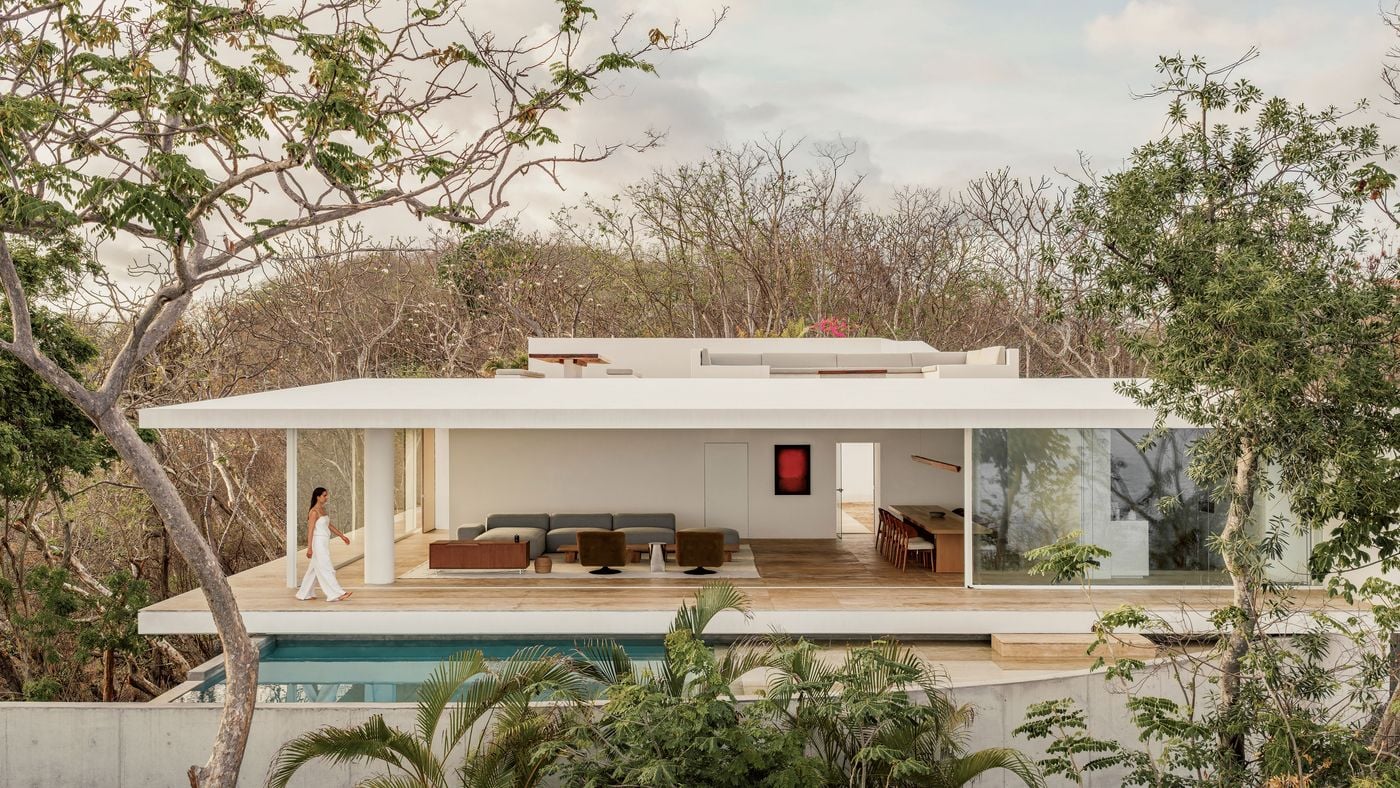 Seen from across the pool, Casa Malonic’s main living level reads as a broad horizontal pavilion suspended among the trees. Retracted glazing opens the lounge and dining areas directly to the terrace, while pale stone flooring, low furniture and the flat white roof reinforce the project’s restrained modernist language. Seen from across the pool, Casa Malonic’s main living level reads as a broad horizontal pavilion suspended among the trees. Retracted glazing opens the lounge and dining areas directly to the terrace, while pale stone flooring, low furniture and the flat white roof reinforce the project’s restrained modernist language.