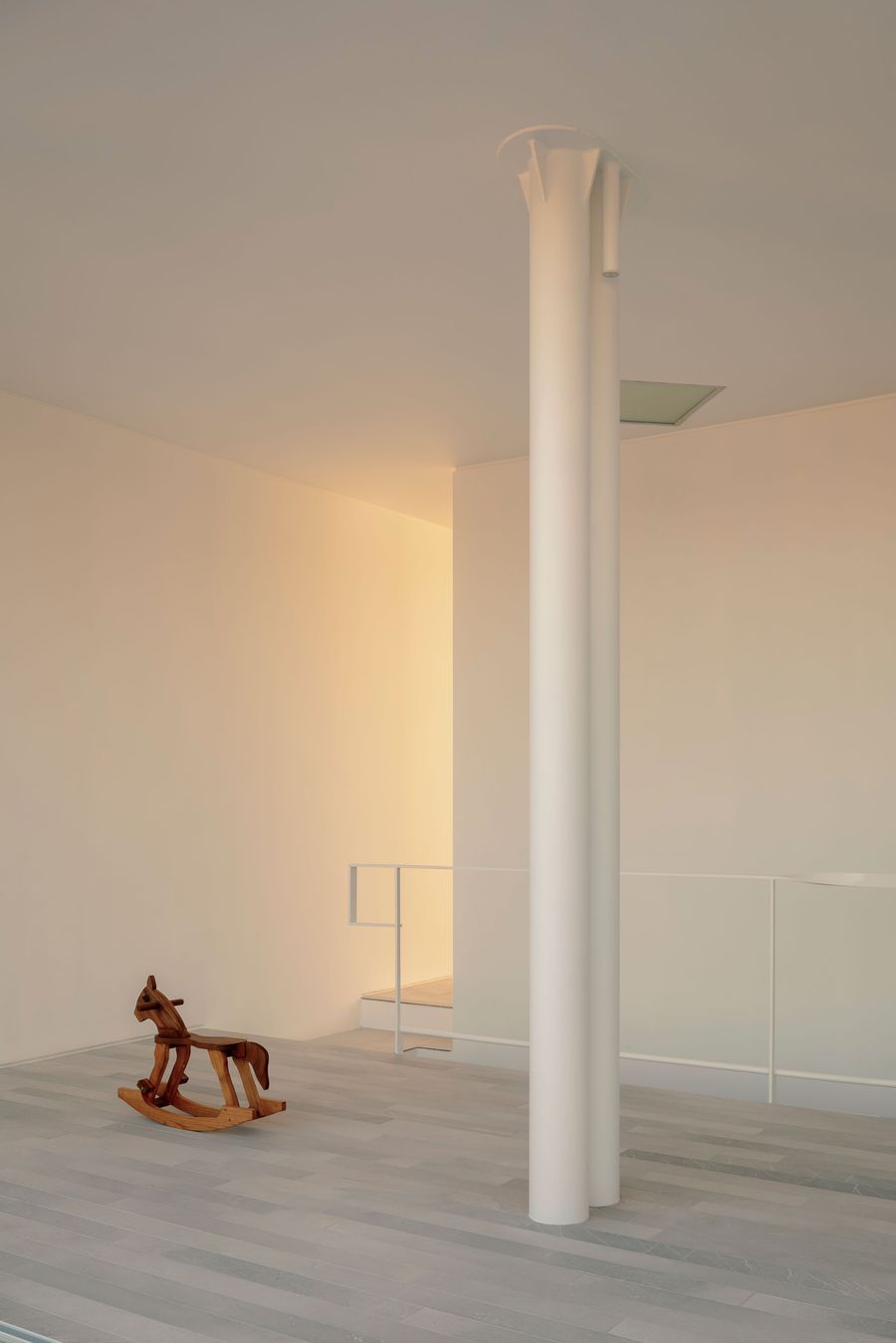On the upper level, a couple of cylindrical white columns rises through an otherwise spare interior of pale stone flooring and softly lit walls. A wooden rocking horse provides a warm, playful counterpoint to the architecture’s severe minimalism, while the stair opening reveals the house’s vertical organisation. On the upper level, a couple of cylindrical white columns rises through an otherwise spare interior of pale stone flooring and softly lit walls. A wooden rocking horse provides a warm, playful counterpoint to the architecture’s severe minimalism, while the stair opening reveals the house’s vertical organisation.