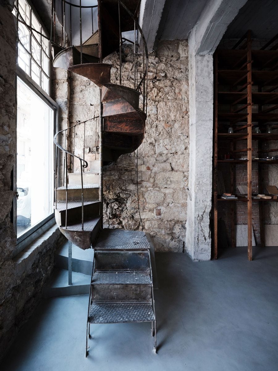 A closer view of the original rusted spiral staircase highlights its patinated steel construction and slender curved balustrade against the rough stone wall. A small freestanding metal stair negotiates the change in level below, while a vintage timber shelving unit occupies the adjoining bay. A closer view of the original rusted spiral staircase highlights its patinated steel construction and slender curved balustrade against the rough stone wall. A small freestanding metal stair negotiates the change in level below, while a vintage timber shelving unit occupies the adjoining bay.
