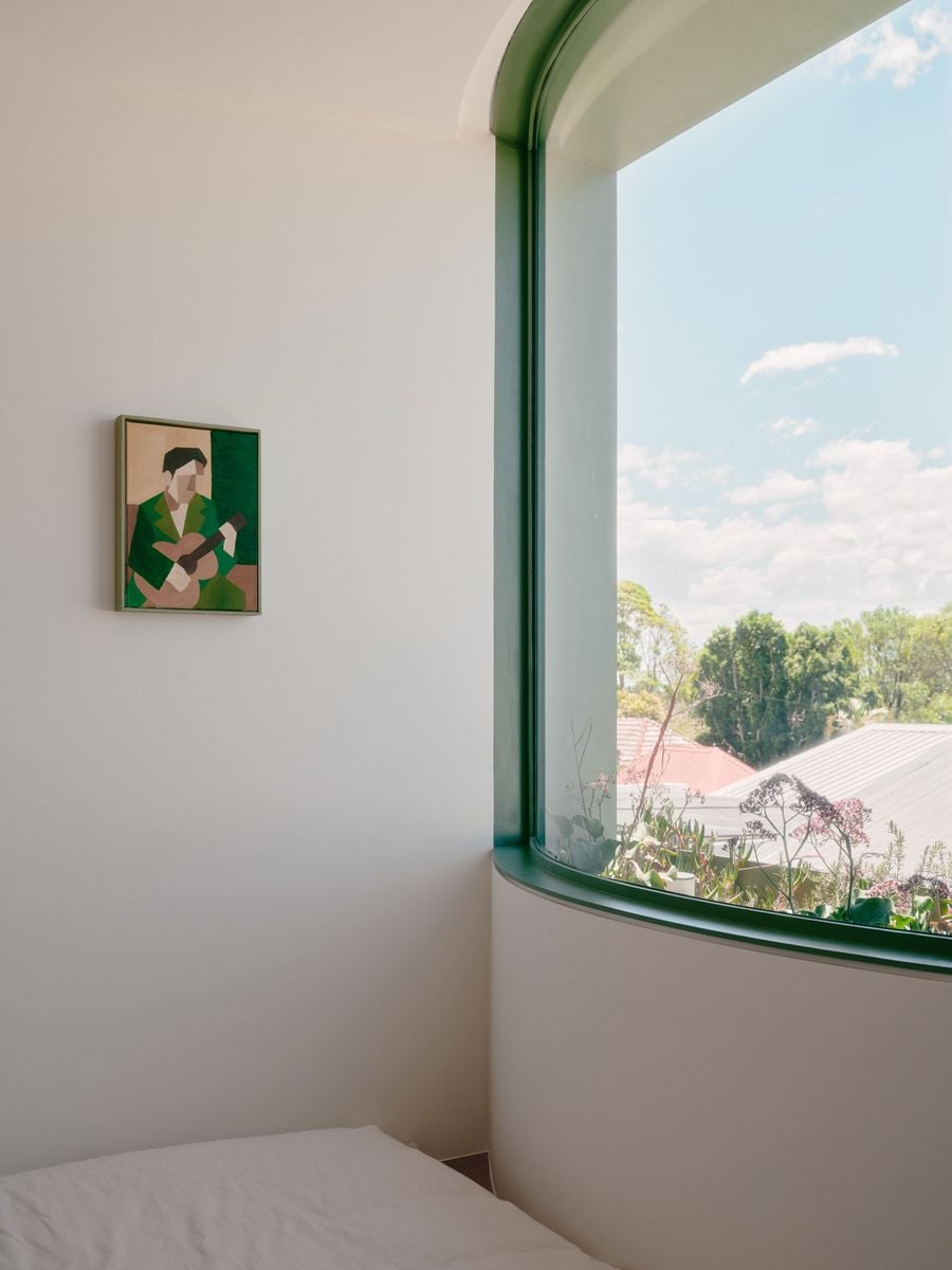 Denj Lane’s bedroom is shaped by a sweeping corner window framed in sage green, its curved sill lined with planting and opening views towards treetops and neighbouring roofs. White walls and bedding keep the compact space calm and minimal, while a small green-toned artwork subtly reinforces the project’s palette. Denj Lane’s bedroom is shaped by a sweeping corner window framed in sage green, its curved sill lined with planting and opening views towards treetops and neighbouring roofs. White walls and bedding keep the compact space calm and minimal, while a small green-toned artwork subtly reinforces the project’s palette.