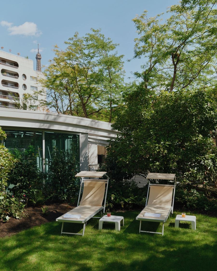 The Ken Club’s solarium garden offers a quiet outdoor counterpoint to the more theatrical interiors. Two cream sun loungers sit among dense planting and mature trees, with the club’s low glazed structure behind and the Eiffel Tower appearing in the distance above the surrounding rooftops. The Ken Club’s solarium garden offers a quiet outdoor counterpoint to the more theatrical interiors. Two cream sun loungers sit among dense planting and mature trees, with the club’s low glazed structure behind and the Eiffel Tower appearing in the distance above the surrounding rooftops.