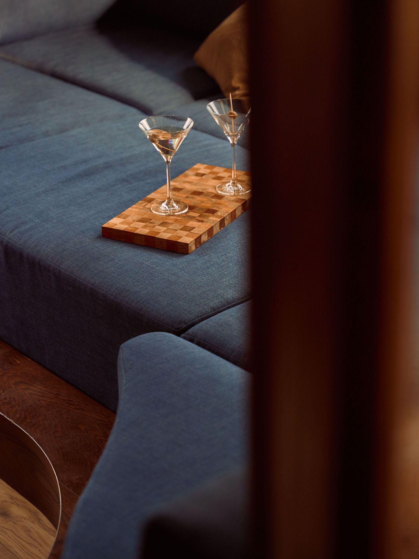 Two martinis rest on a chequered timber tray within the room’s deep-blue sofa pit, reinforcing Holloway Li’s playful nod to 1960s after-hours glamour. The close crop foregrounds the upholstery’s generous scale and the social character of a feature conceived as more than conventional hotel seating. Two martinis rest on a chequered timber tray within the room’s deep-blue sofa pit, reinforcing Holloway Li’s playful nod to 1960s after-hours glamour. The close crop foregrounds the upholstery’s generous scale and the social character of a feature conceived as more than conventional hotel seating.