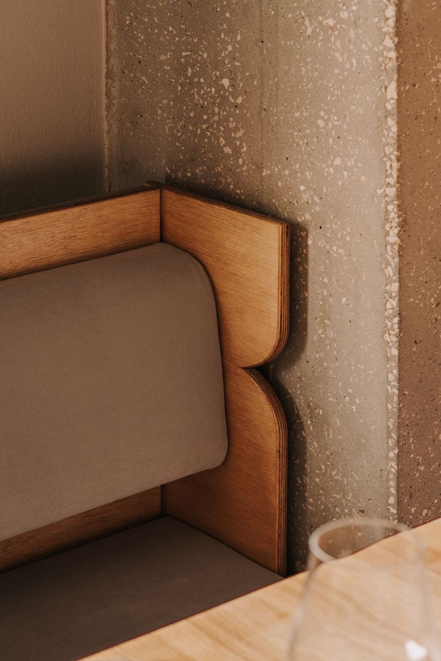 A detail of the upstairs dining banquette at WIN wine bar in Warsaw reveals its sculptural plywood frame, whose curved, layered profile embraces soft taupe upholstery. Set against an exposed aggregate concrete wall, the warm timber introduces a tactile contrast to the building’s raw industrial surfaces. A detail of the upstairs dining banquette at WIN wine bar in Warsaw reveals its sculptural plywood frame, whose curved, layered profile embraces soft taupe upholstery. Set against an exposed aggregate concrete wall, the warm timber introduces a tactile contrast to the building’s raw industrial surfaces.