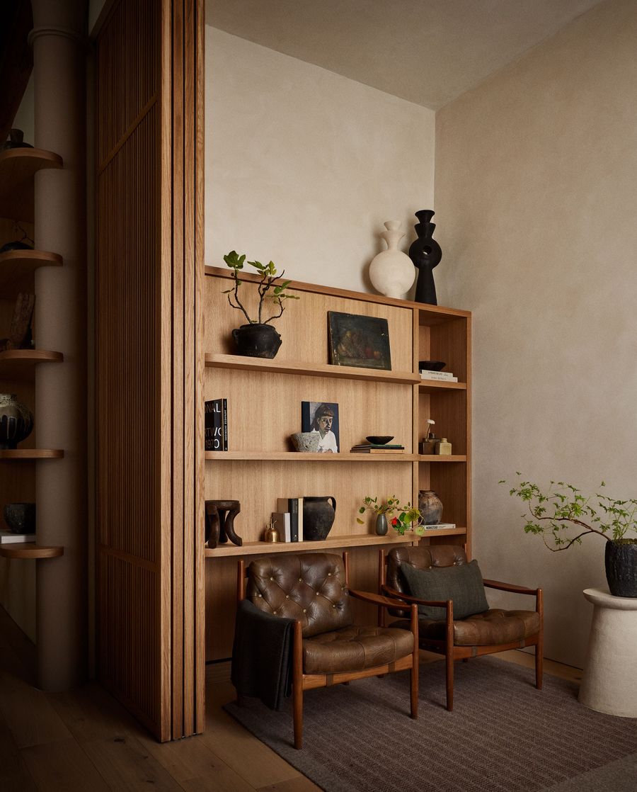A cozy corner featuring two vintage chairs, a wooden shelf with decorative vases, books, and a small potted plant on a table. A cozy corner featuring two vintage chairs, a wooden shelf with decorative vases, books, and a small potted plant on a table.