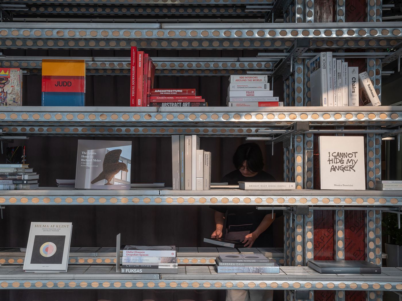 Within the sophisticated industrial framework of 889 GLO Art Space, a person engages with books displayed on the modular shelving system. The shelves, constructed from perforated metal, hold a curated collection of art and design books, reflecting the space's commitment to culture and its integration of a raw, functional aesthetic with intellectual pursuits. Within the sophisticated industrial framework of 889 GLO Art Space, a person engages with books displayed on the modular shelving system. The shelves, constructed from perforated metal, hold a curated collection of art and design books, reflecting the space's commitment to culture and its integration of a raw, functional aesthetic with intellectual pursuits.