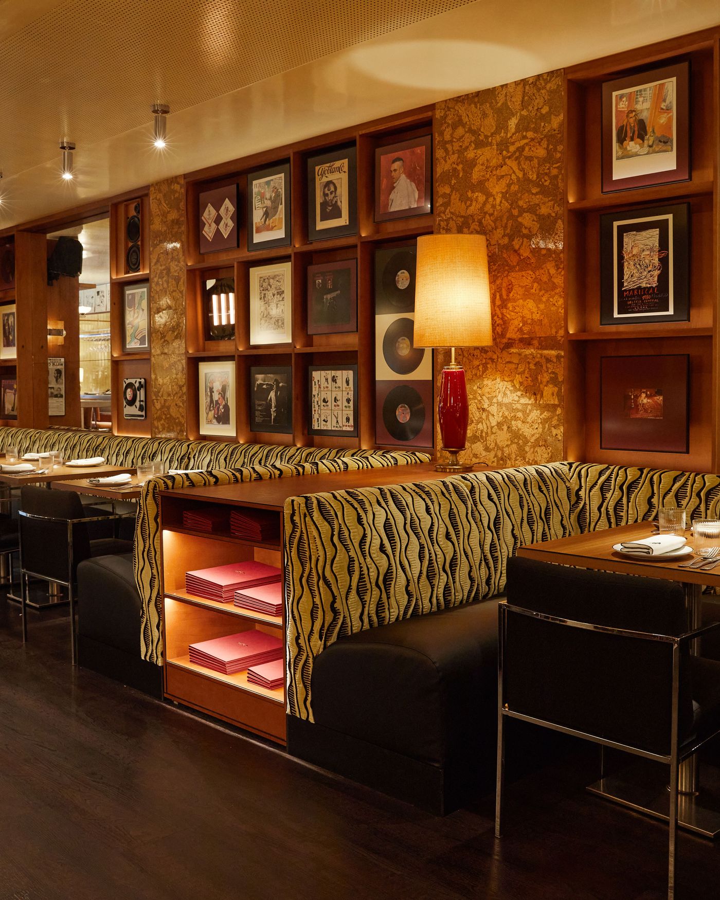 This view of the dining area highlights a maximalist, vintage aesthetic. The space is filled with a bold, black and yellow patterned banquette that incorporates shelving for menus. A vibrant red lamp on a table adds a pop of color, complementing the extensive gallery wall with its collection of framed records and art. This view of the dining area highlights a maximalist, vintage aesthetic. The space is filled with a bold, black and yellow patterned banquette that incorporates shelving for menus. A vibrant red lamp on a table adds a pop of color, complementing the extensive gallery wall with its collection of framed records and art.